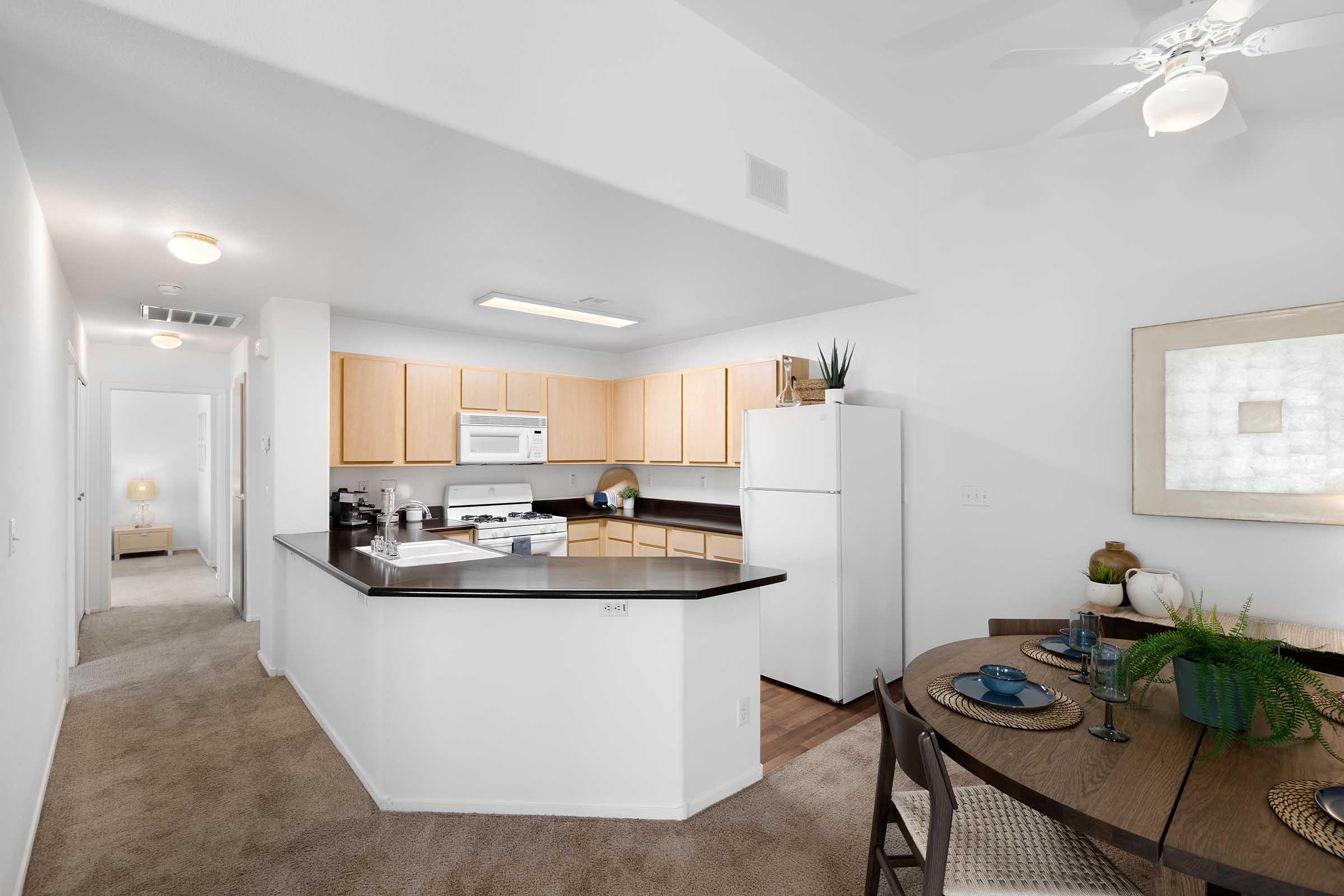 A modern kitchen area with light wooden cabinets, a black countertop, and white appliances, including a stove and refrigerator. A small dining table with decorative items and plants is set nearby. The space features beige carpeting and a ceiling fan. A hallway can be seen in the background.