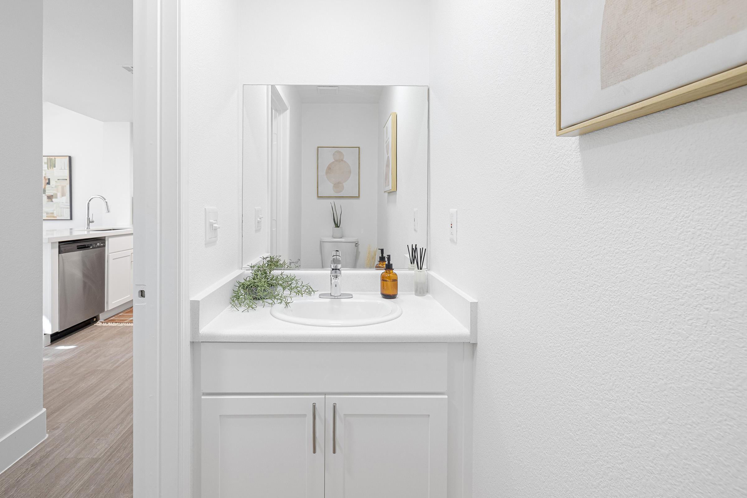 A well-lit, modern bathroom featuring a white sink with a mirror above it. The counter displays a small plant and decorative bottles. The walls are painted white, and there is a framed abstract artwork nearby. In the background, a glimpse of a kitchen area is visible, showcasing contemporary design elements.