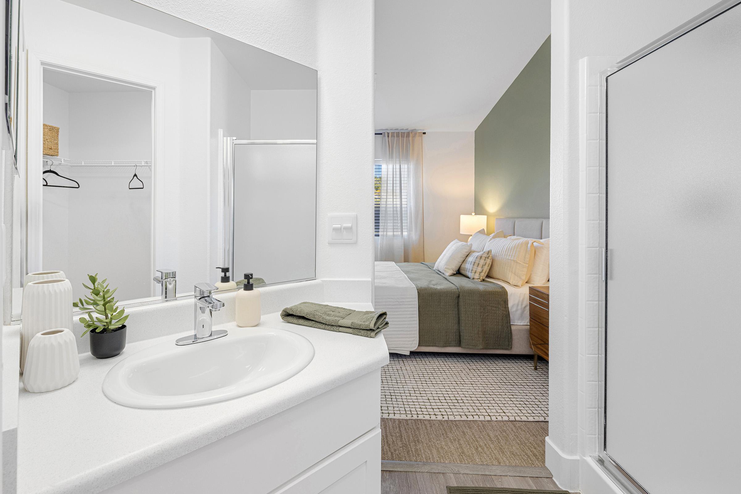A well-lit bathroom featuring a white vanity with a sink, decorative plant, soap dispenser, and towels. In the background, a cozy bedroom is visible with a bed dressed in neutral tones and natural light streaming through a window. The overall decor is modern and inviting.