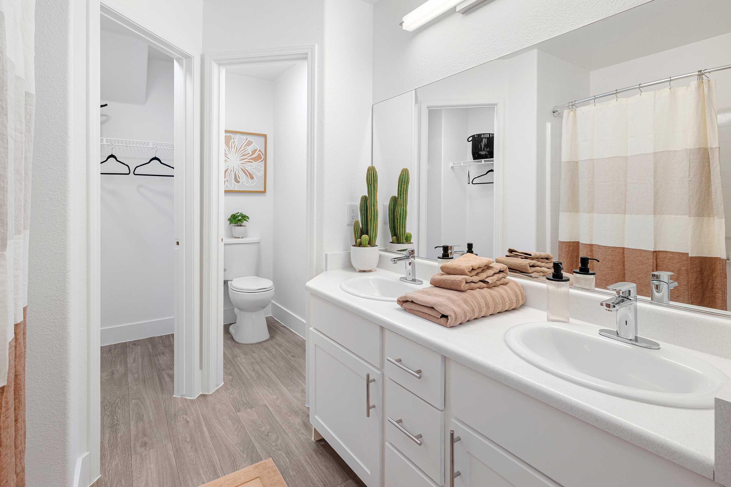 A modern bathroom featuring a double sink with a white countertop, neatly arranged towels in light brown tones, and decorative plants. The space is well-lit, with a mirror above the sinks and a shower curtain in soft stripes. A toilet and hooks for towels are visible in the background.
