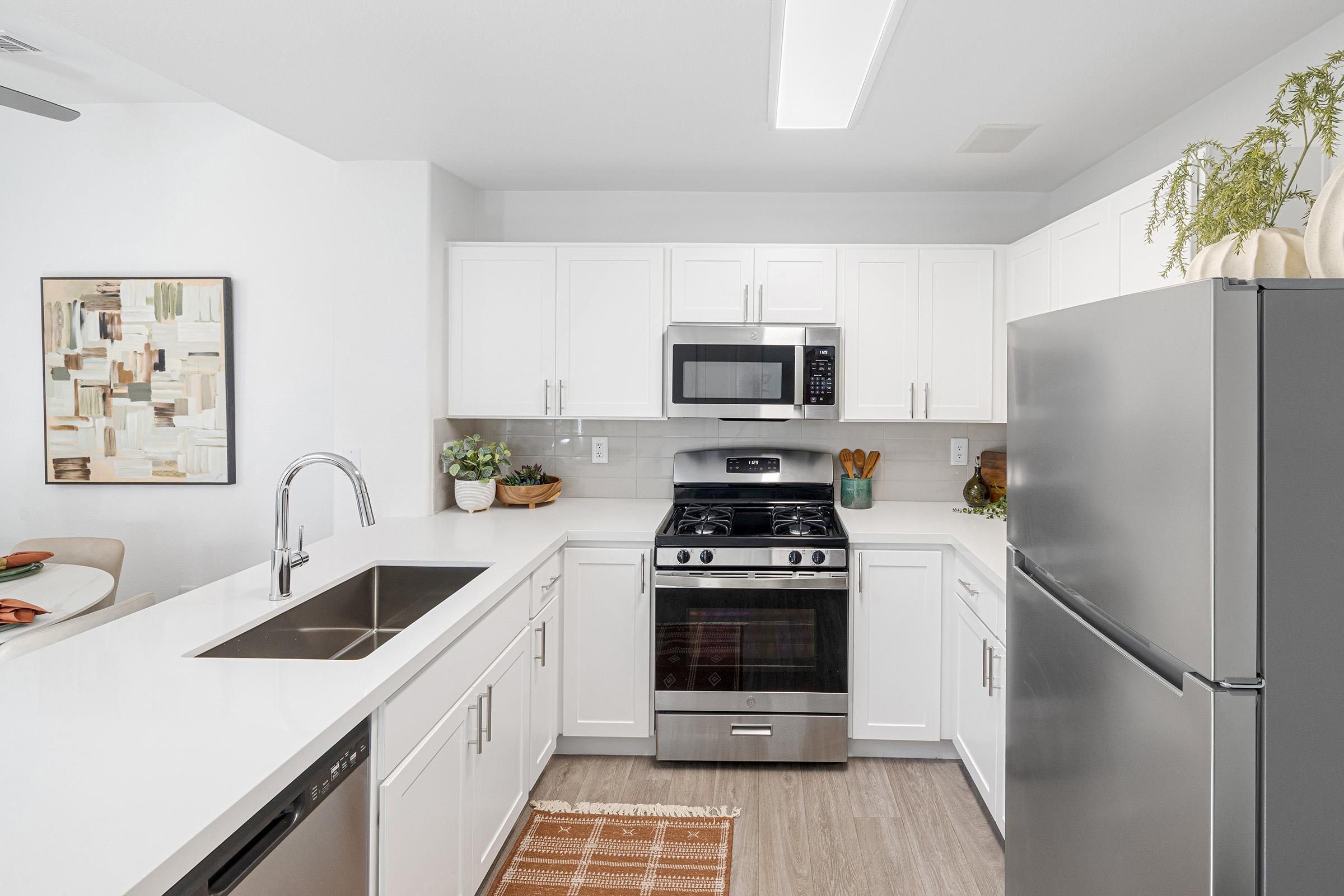 Modern kitchen featuring white cabinets, stainless steel appliances, and a sleek countertop. It includes a gas stove, built-in microwave, and a double sink. Decorative plants and a framed artwork add a touch of style. The flooring is light wood, creating a bright and inviting atmosphere.