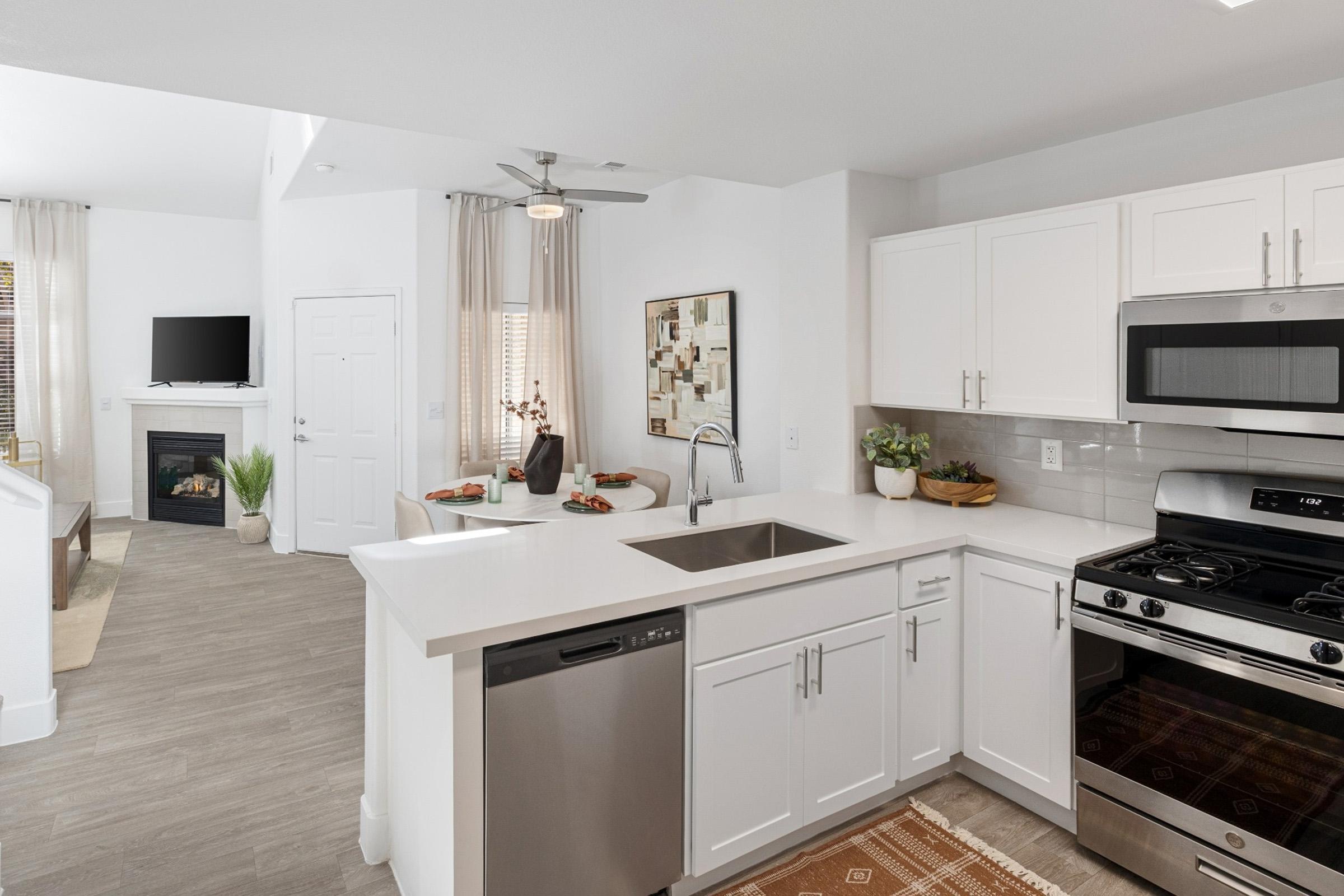 Modern kitchen featuring white cabinetry, a stainless steel sink and stove, and a dishwasher. Complemented by a light countertop and a decorative rug, the open layout connects to a dining area with a small table and chairs, and includes a cozy living space with a fireplace and TV in the background.