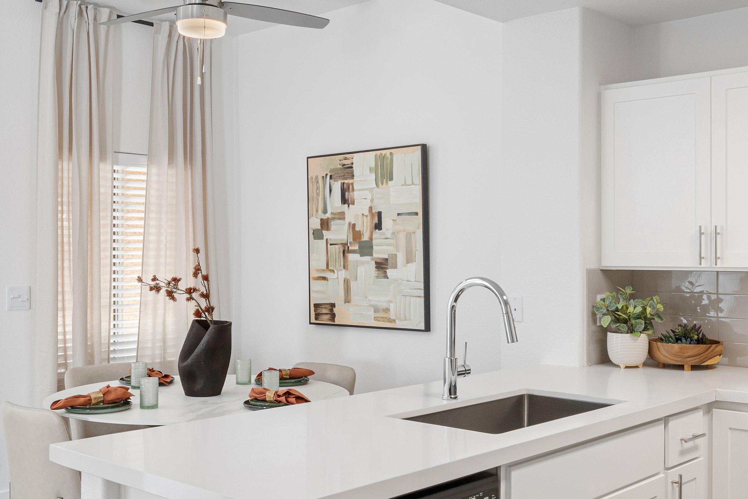 A modern kitchen scene featuring a white countertop and sink. In the background, there’s a dining area with a table set for two, adorned with green plates and cloth napkins. A large abstract painting hangs on the wall, and plants are placed on the counter and table, enhancing the cozy atmosphere. Bright, natural light filters through the window.