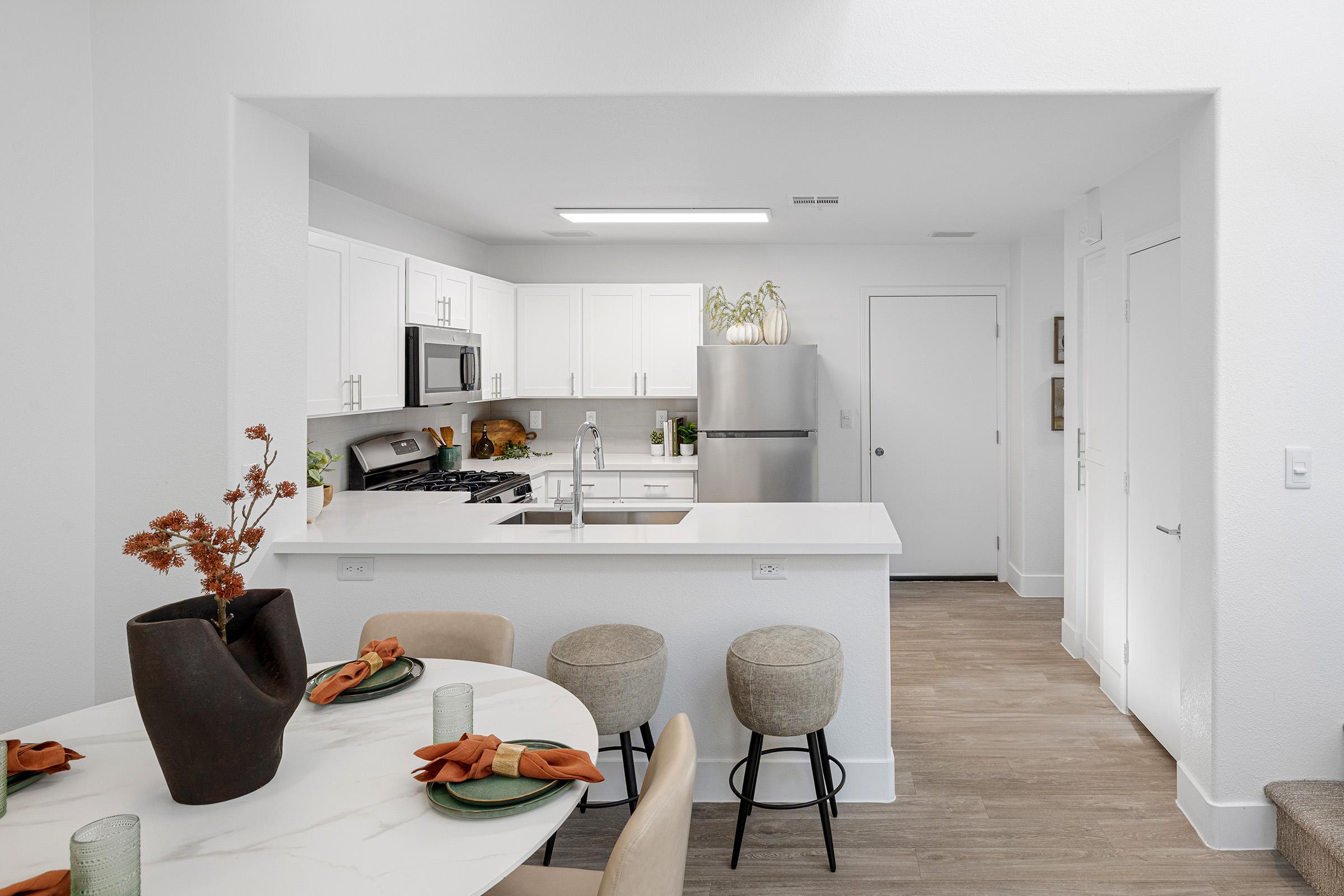 A modern kitchen with white cabinets, stainless steel appliances, and a large island. In the foreground, a dining table is set with green plates and orange napkins, accompanied by two beige bar stools. Natural light fills the space, highlighting the clean design and minimalist decor.