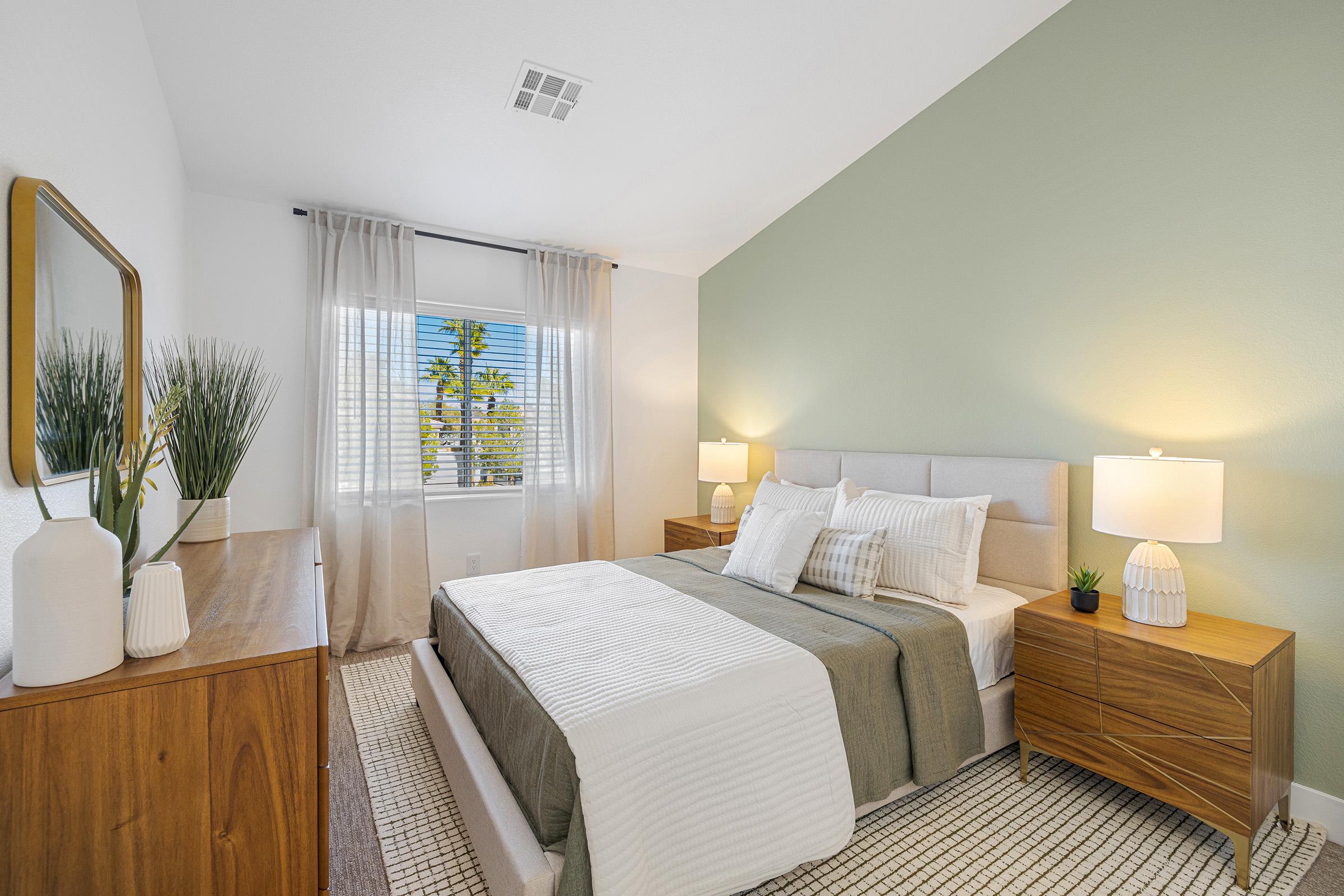 A modern bedroom featuring a comfortable queen-sized bed with neutral bedding and decorative pillows. Light wooden nightstands with lamps are on either side of the bed. A dresser stands against the wall, and a window with sheer curtains offers natural light, revealing greenery outside.