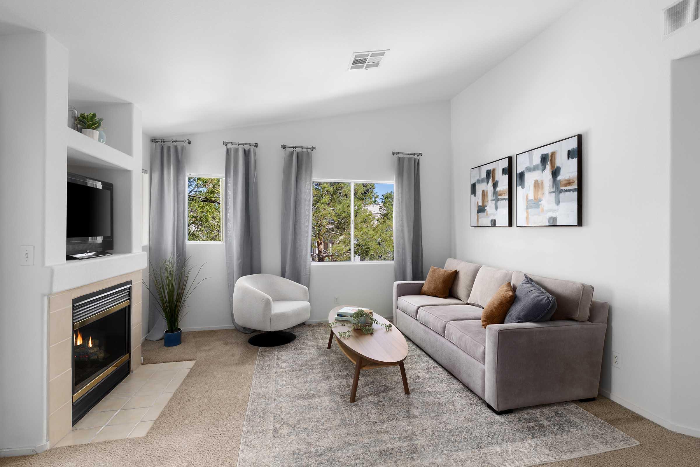A cozy living room featuring a gray sofa with decorative pillows, a modern white accent chair, a round coffee table, and a fireplace. Large windows with light curtains allow natural light in, and artwork is displayed on the wall. The space is designed with neutral colors and a comfortable, inviting atmosphere.