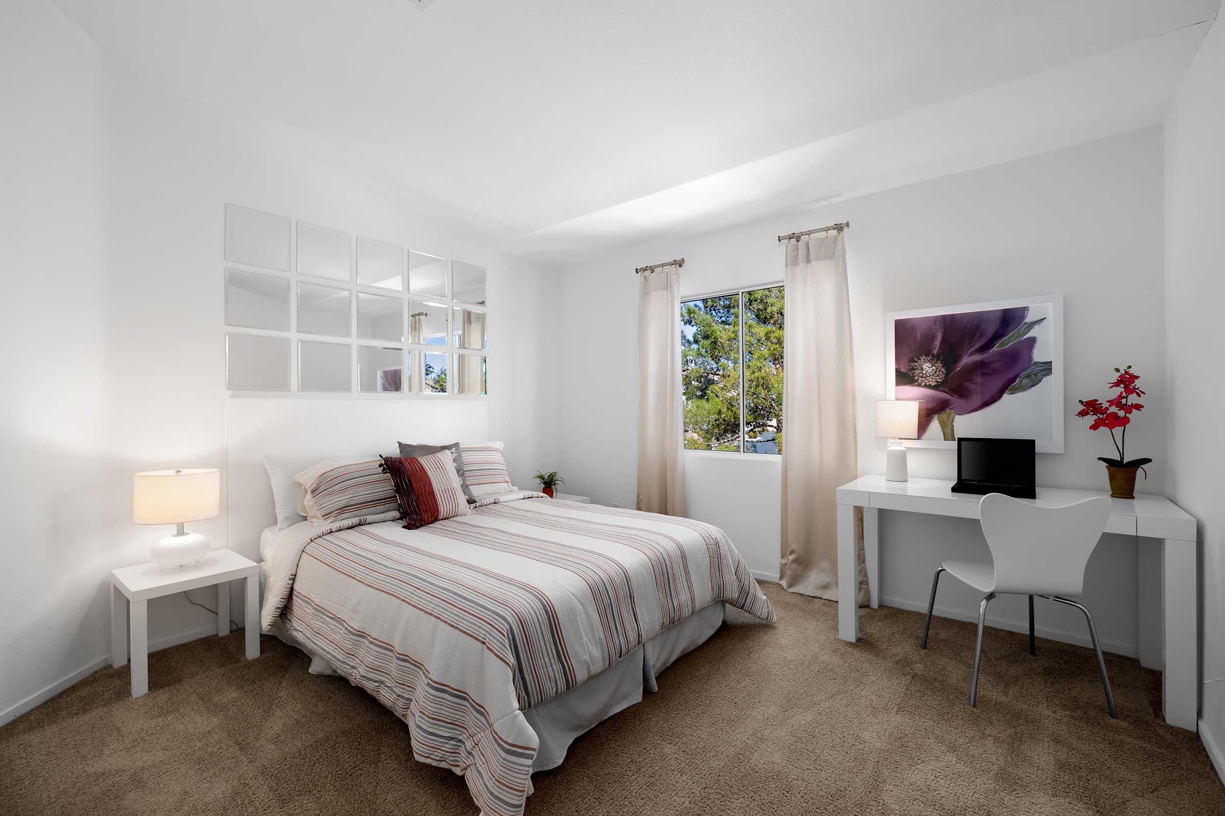 A modern bedroom featuring a striped bedspread on a double bed, a white desk with a computer and chair, a small nightstand with a lamp, and a window with sheer curtains. A floral artwork hangs on the wall, and a potted plant adds a touch of greenery. The room has a neutral color scheme and soft carpet.