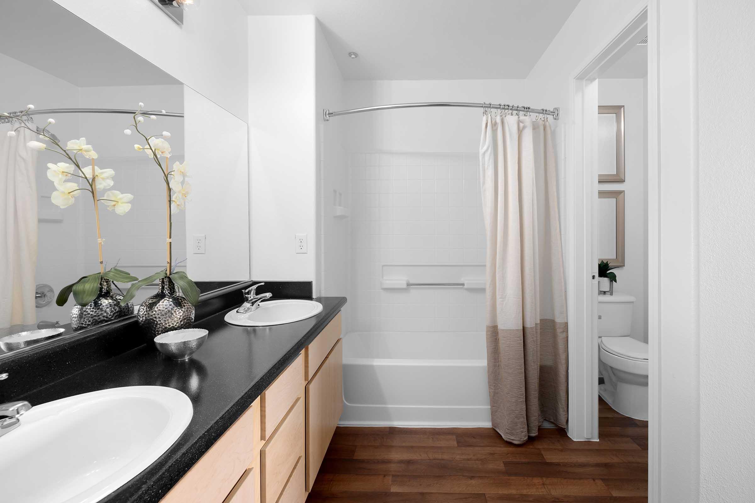 A modern bathroom featuring a double vanity with a dark countertop, two sinks, and decorative elements like potted orchids. The space includes a shower/tub combo with a light-colored curtain, wood flooring, and a neutral color scheme. The background shows a toilet in a separate area.