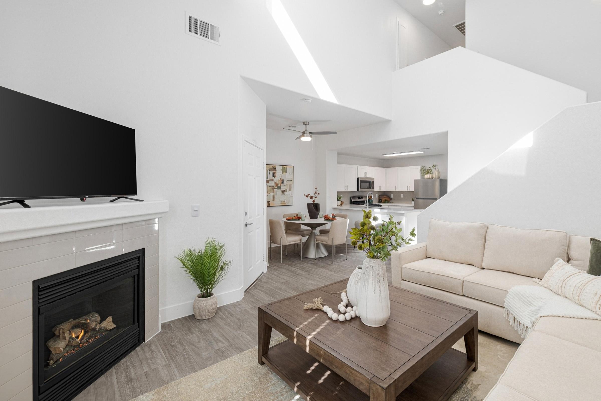 A modern living room with a light-colored sectional sofa and a wooden coffee table adorned with decorative items. A wall-mounted TV is above a stylish fireplace. In the background, a dining area is visible, featuring a round table with chairs and a kitchen area with stainless steel appliances. Natural light illuminates the space.
