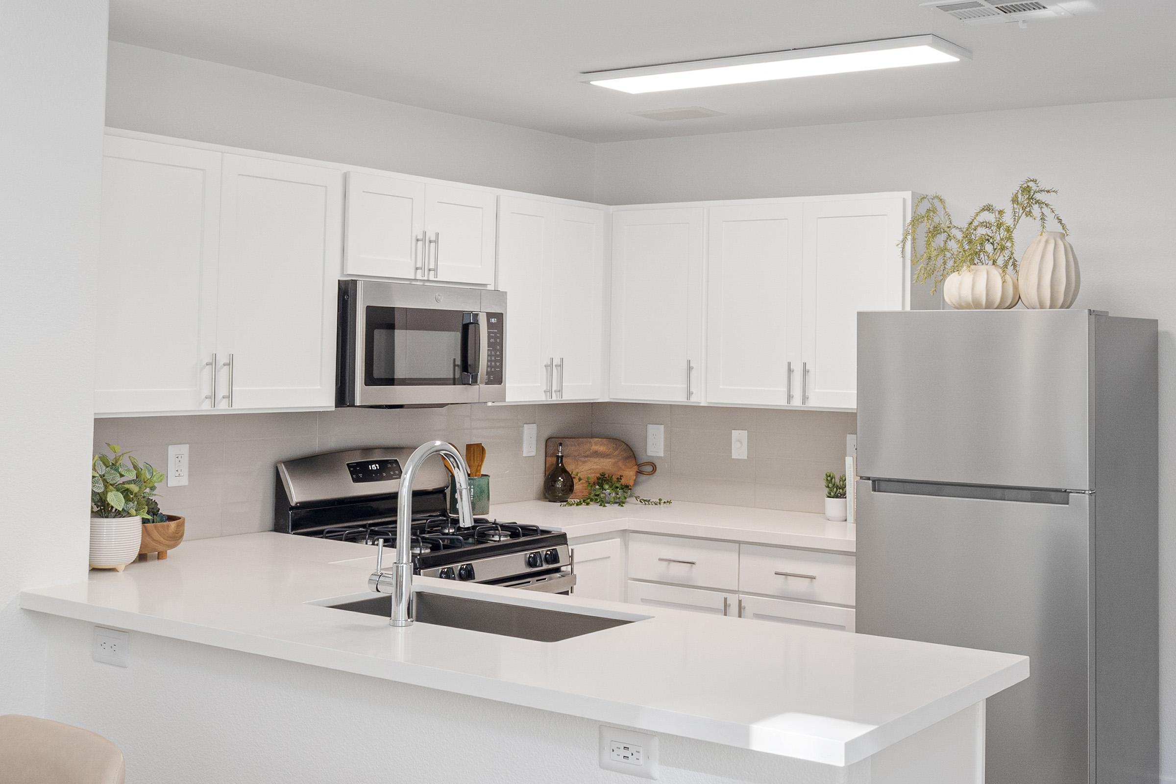 Modern kitchen featuring white cabinetry, stainless steel appliances, and a light-colored countertop. The kitchen includes a gas stove, microwave, and refrigerator, along with decorative plants and a wooden cutting board on the countertop, creating a clean and inviting atmosphere.