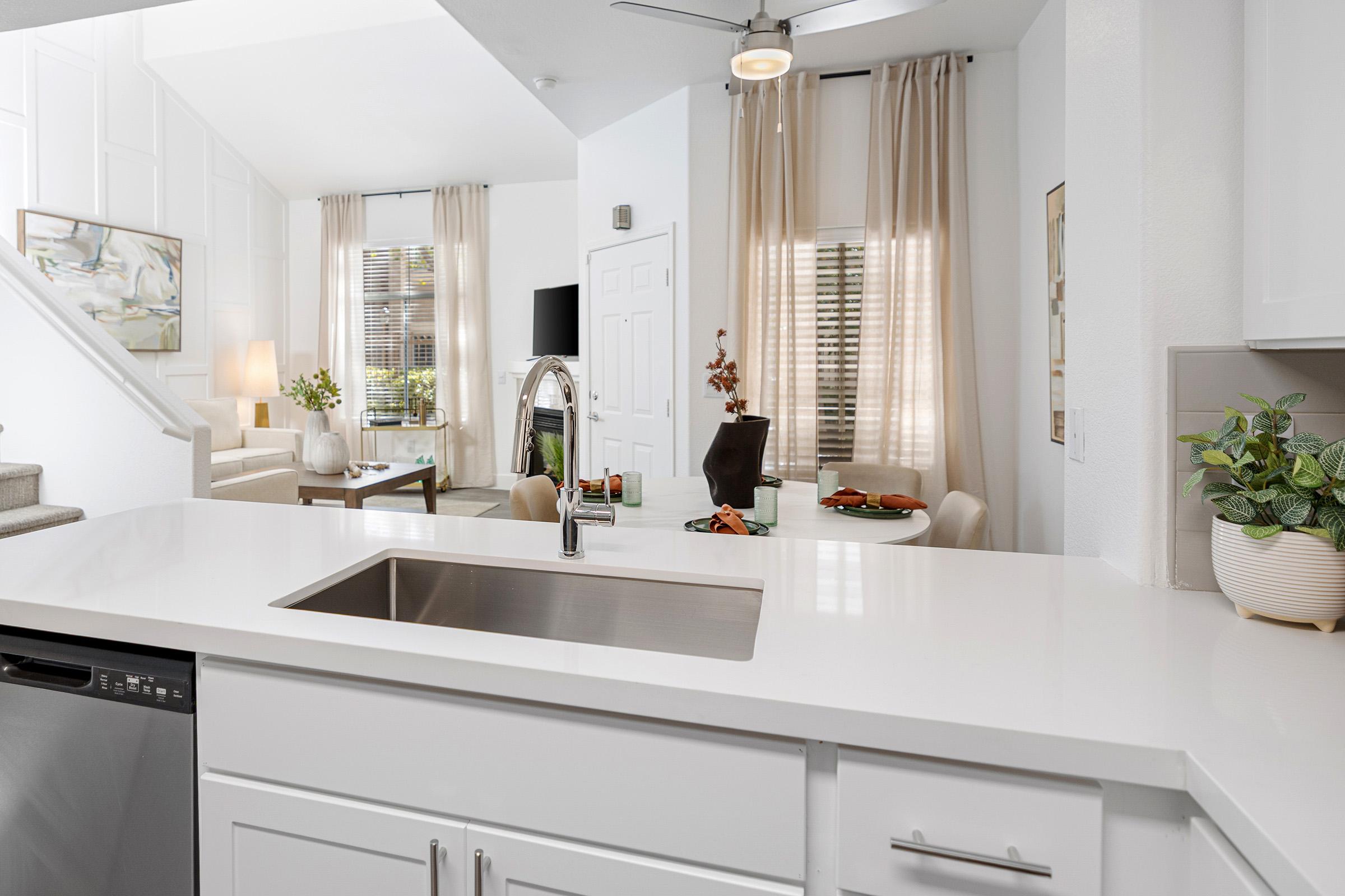 A modern kitchen with a white countertop and stainless steel sink, featuring a view into a stylish living area. Light floods in through large windows with soft curtains. In the background, there is a dining table set for a meal, and a comfortable seating area with a television and decorative plants.