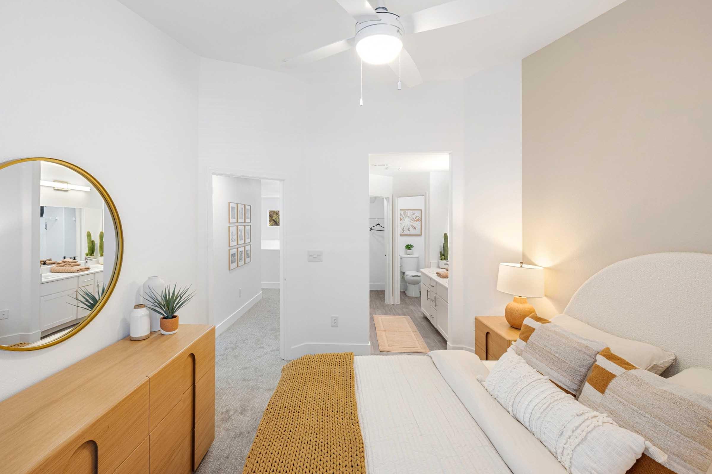 A cozy bedroom featuring a light-colored wall, a large bed with decorative pillows, and a textured throw. There's a dresser, a round mirror on the wall, and a small plant. To the left, a doorway leads to a separate room, and to the right, a doorway reveals a bathroom space. A ceiling fan adds to the ambiance.