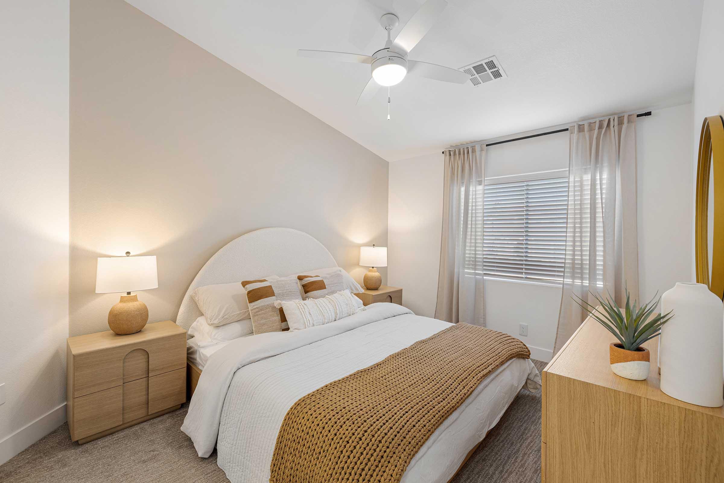 Cozy bedroom featuring a bed with a neutral-colored comforter, accent pillows, and a stylish headboard. Two bedside tables with lamps flank the bed. Natural light filters through a window with sheer curtains, and a minimalist decor is complemented by a wooden dresser and a potted plant.