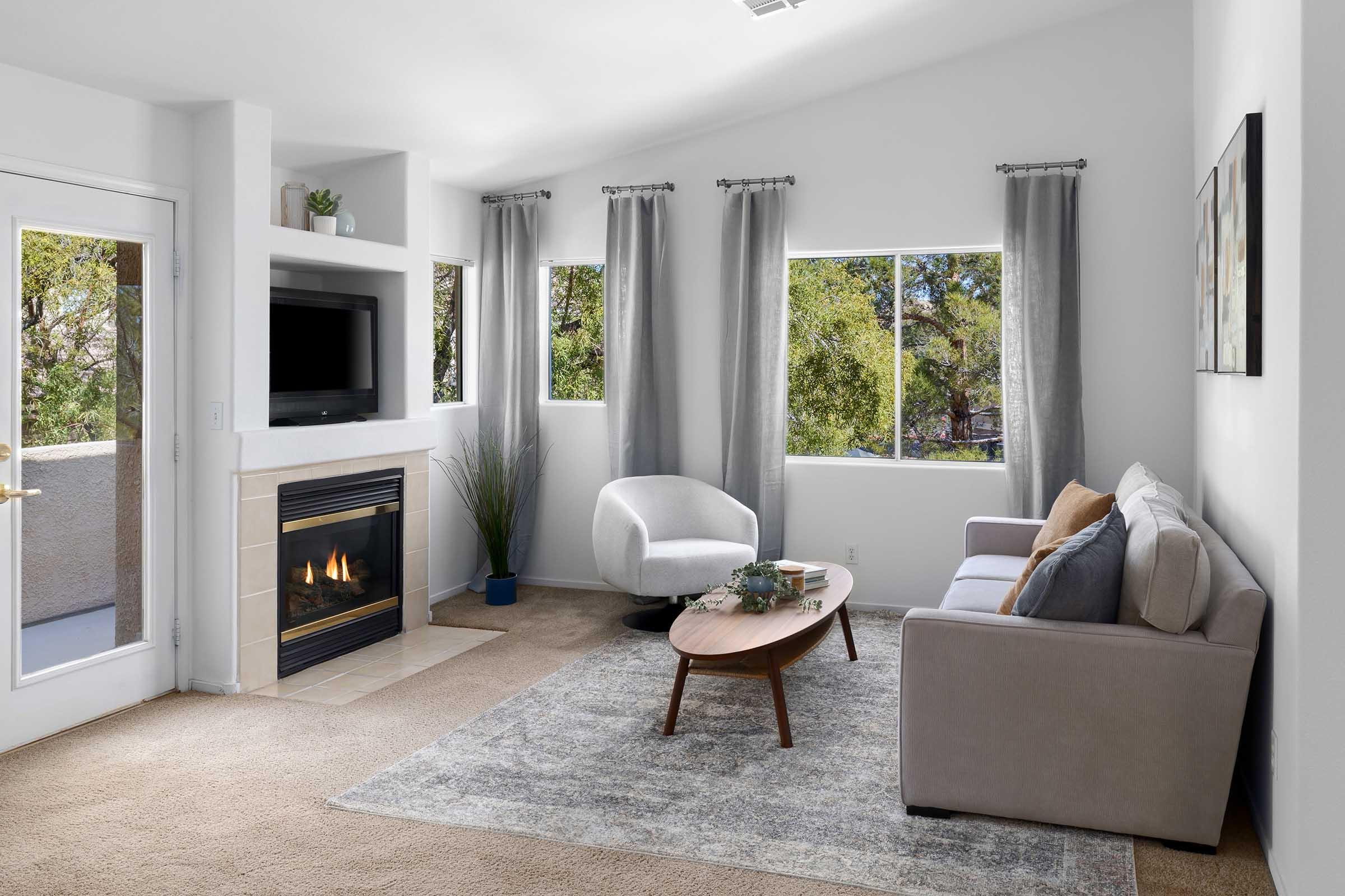 A cozy living room featuring a beige sofa and a round wooden coffee table on a light rug. There's a modern fireplace built into the wall, with a TV mounted above it. Large windows allow natural light to enter, adorned with light gray curtains. Green trees are visible through the windows, enhancing the serene atmosphere.