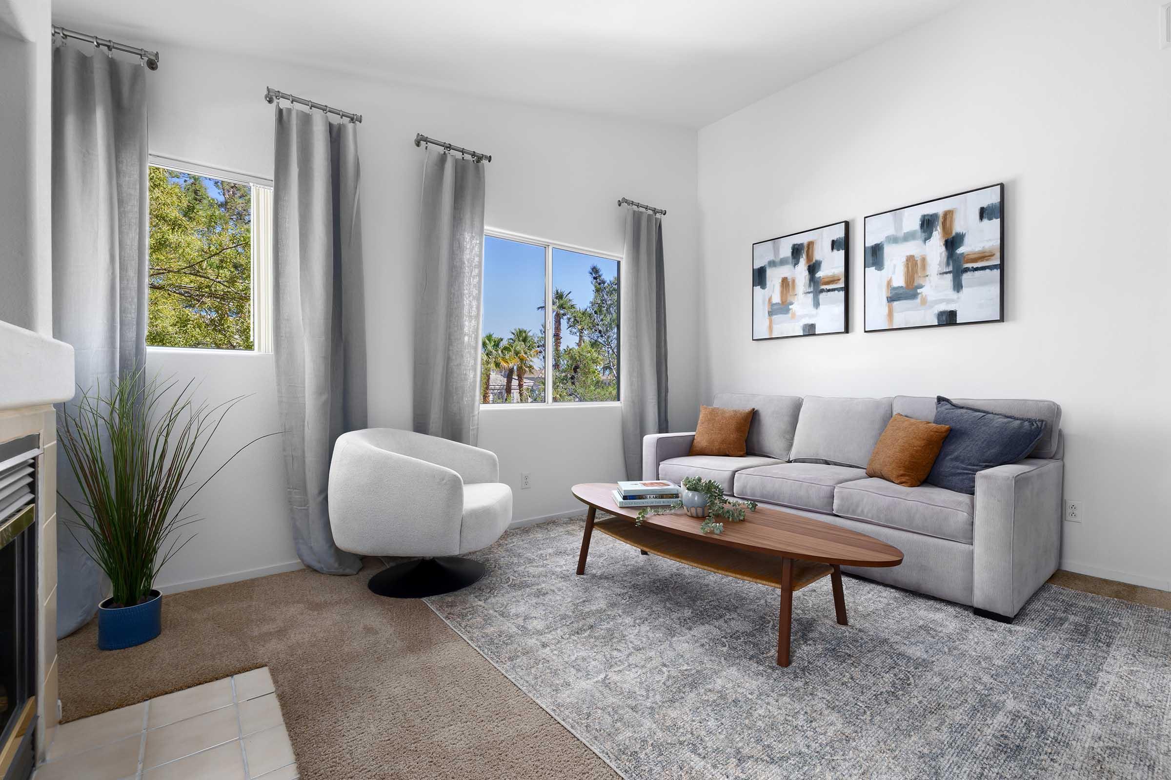 A modern living room featuring a gray sofa with blue and orange pillows, a stylish circular chair, a wooden coffee table with decorative items, and a light-colored area rug. Large windows allow natural light to fill the space, framed by sheer curtains, with greenery visible outside.