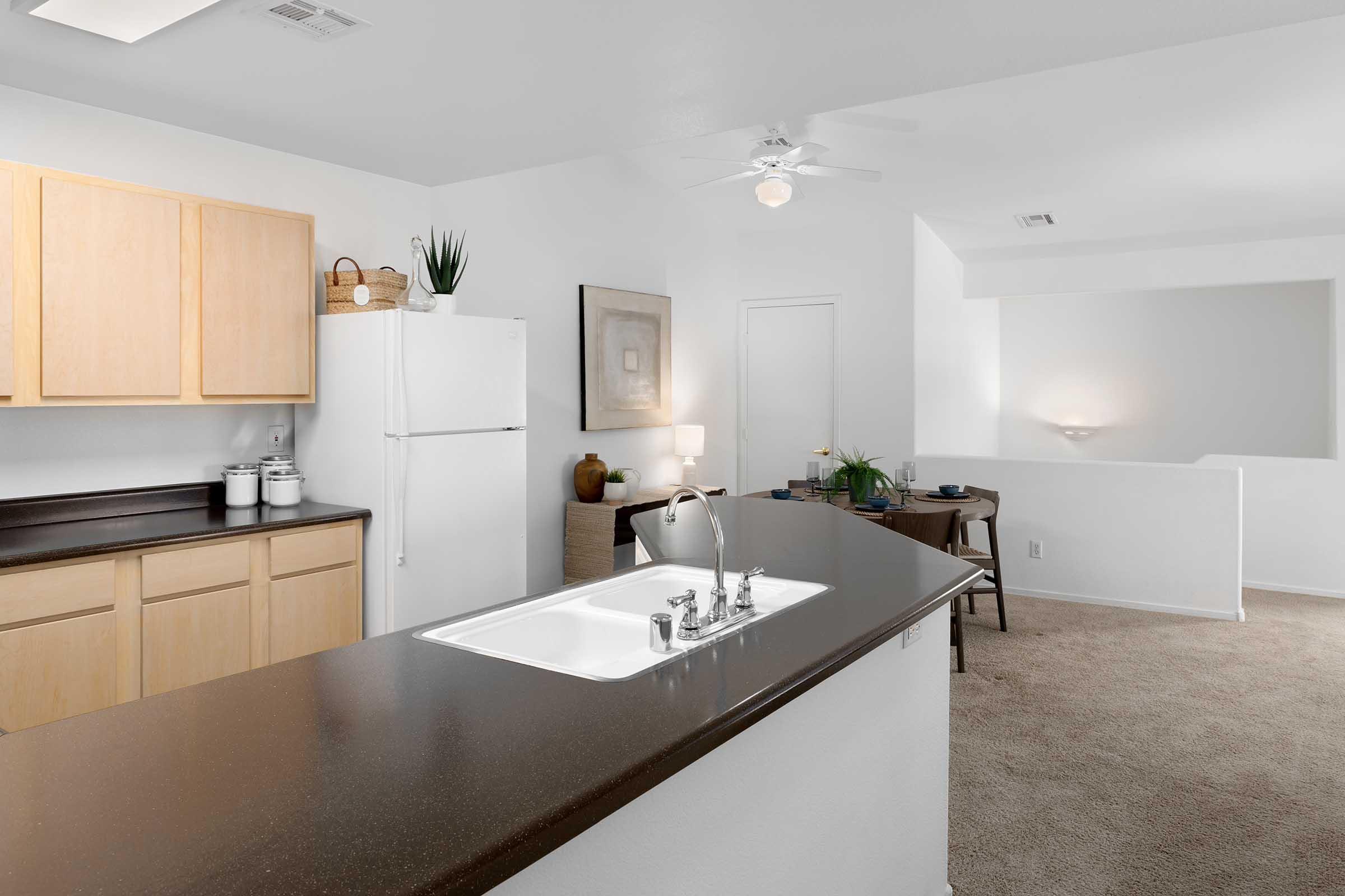 A spacious kitchen featuring light wooden cabinets, a white refrigerator, and a dark countertop. A sink is integrated into the island, with a ceiling fan above. The area is well-lit, with a dining space visible in the background, showcasing a table and decorative plants. The room has neutral wall colors and carpeted flooring.