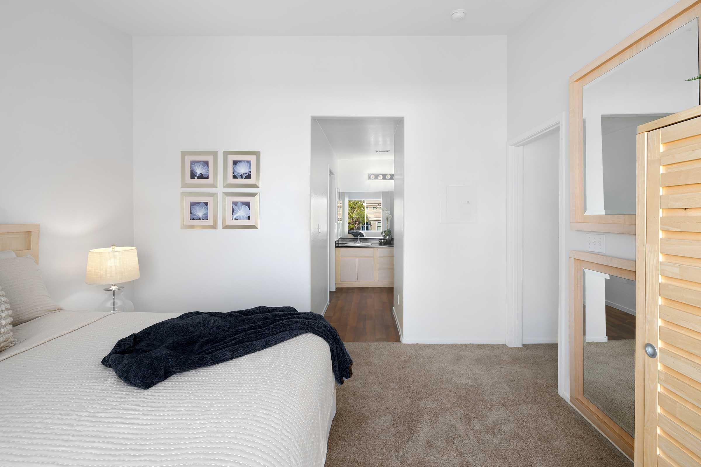 A well-lit bedroom with a bed featuring a textured white blanket. Beside it, a lamp illuminates the space. On the wall, there are three framed pictures. A doorway leads to a kitchen area visible in the background. A wooden cabinet with a mirrored front is positioned next to the bed. The carpet is a soft beige color.