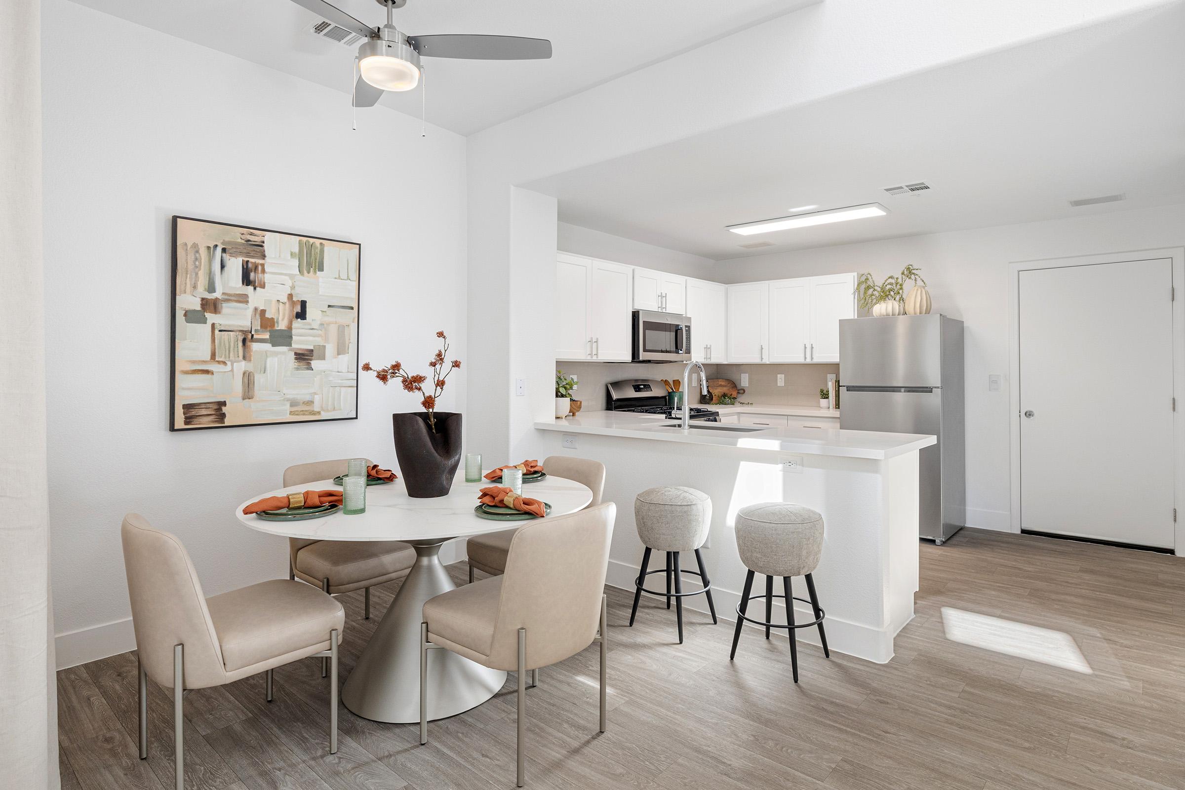 A modern kitchen with an open layout features a round dining table set for four, light-colored chairs, and a painting on the wall. The kitchen includes white cabinets, stainless steel appliances, and a bar seating area with three stools. The space is bright with natural light and has a minimalist design.