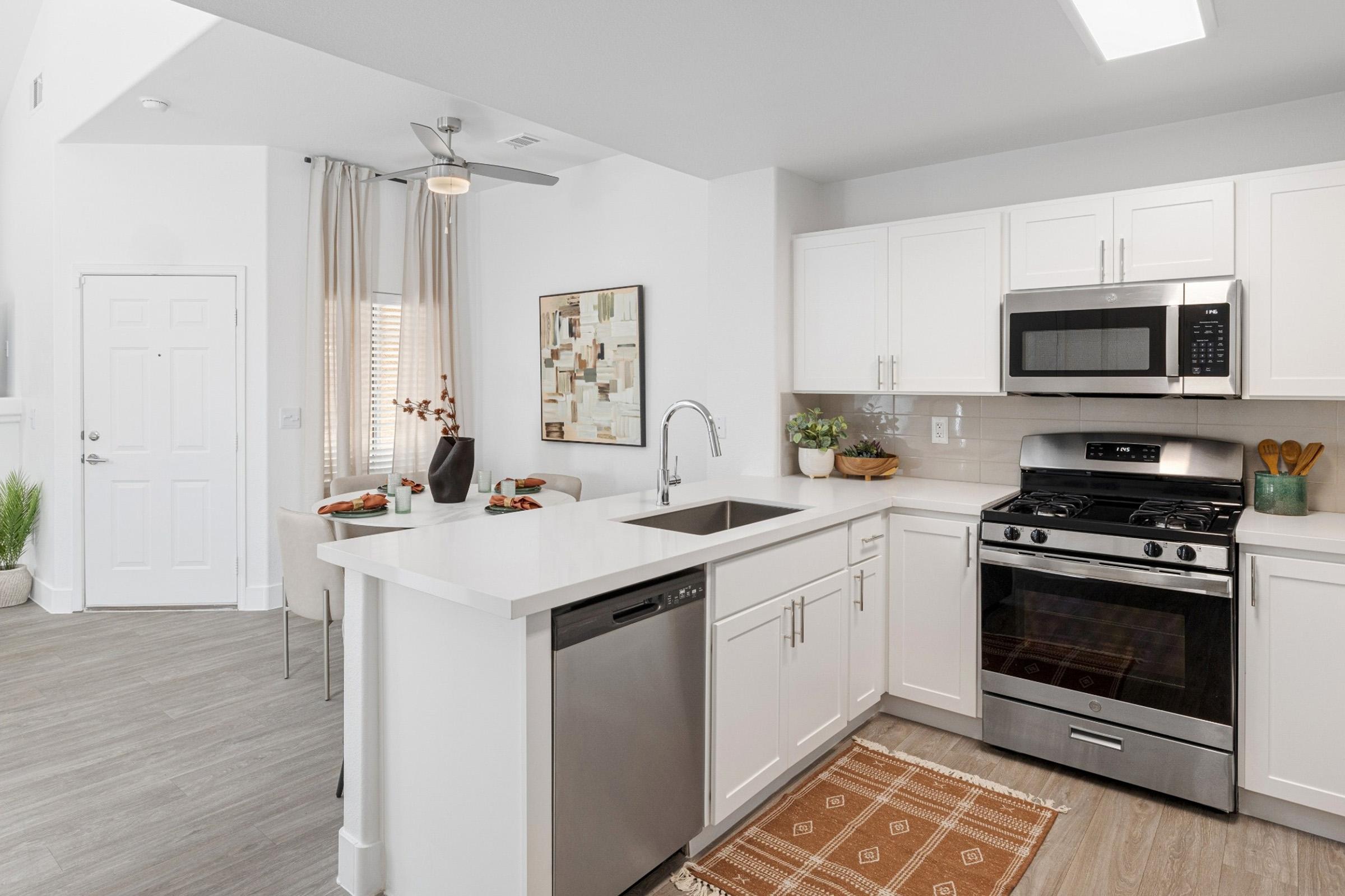 Modern kitchen featuring white cabinetry, a stainless steel refrigerator, oven, and dishwasher. A sleek countertop with a sink is complemented by a decorative rug. Natural light filters in through a window, and a dining area with a table and chairs is visible in the background.