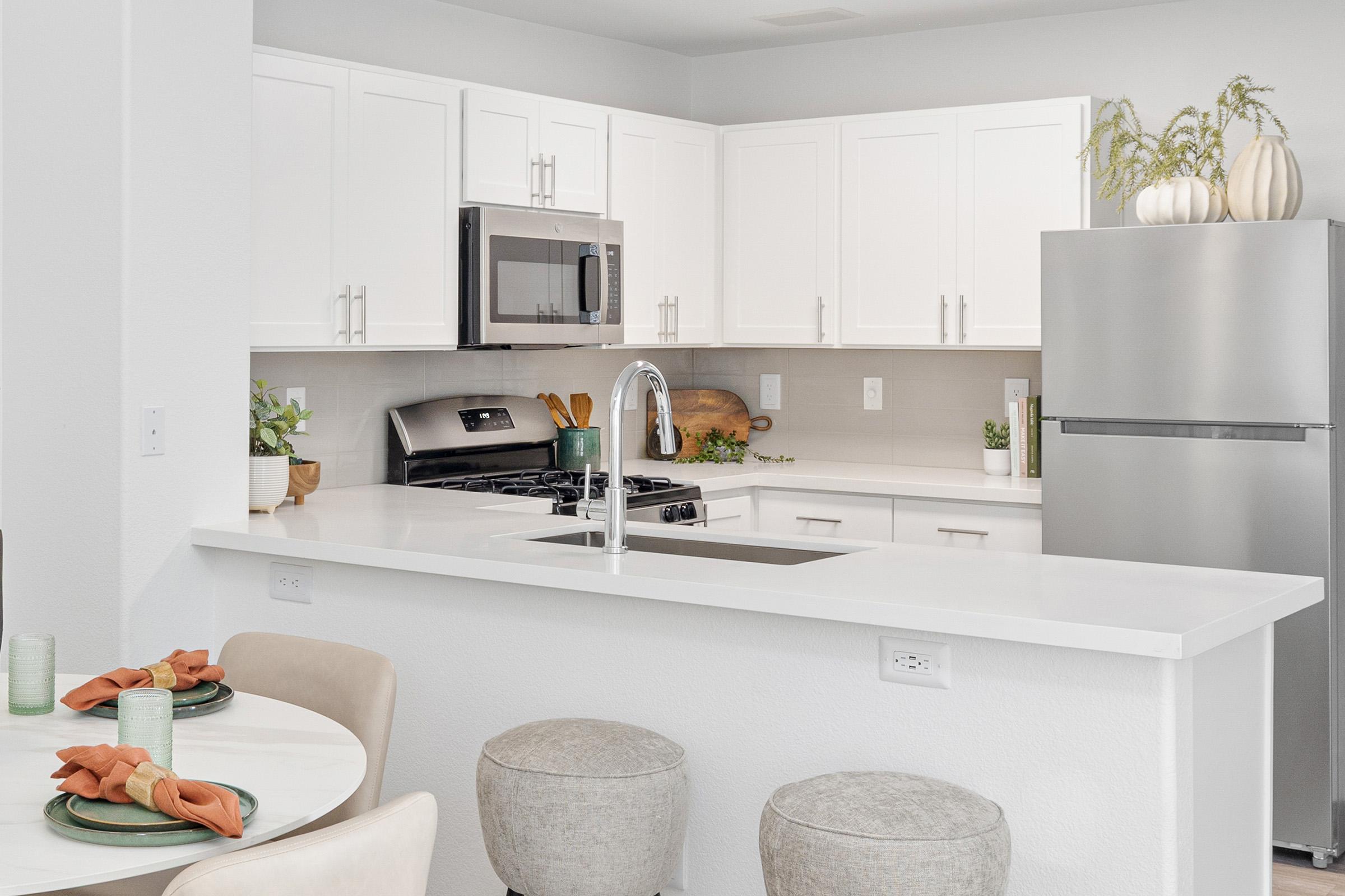 Modern kitchen featuring white cabinetry, stainless steel appliances, and a large island with a sleek countertop. The space includes a dining table set with green glasses and orange napkins, complemented by subtle decorative plants and items on the counter. Soft, neutral tones create a bright, inviting atmosphere.