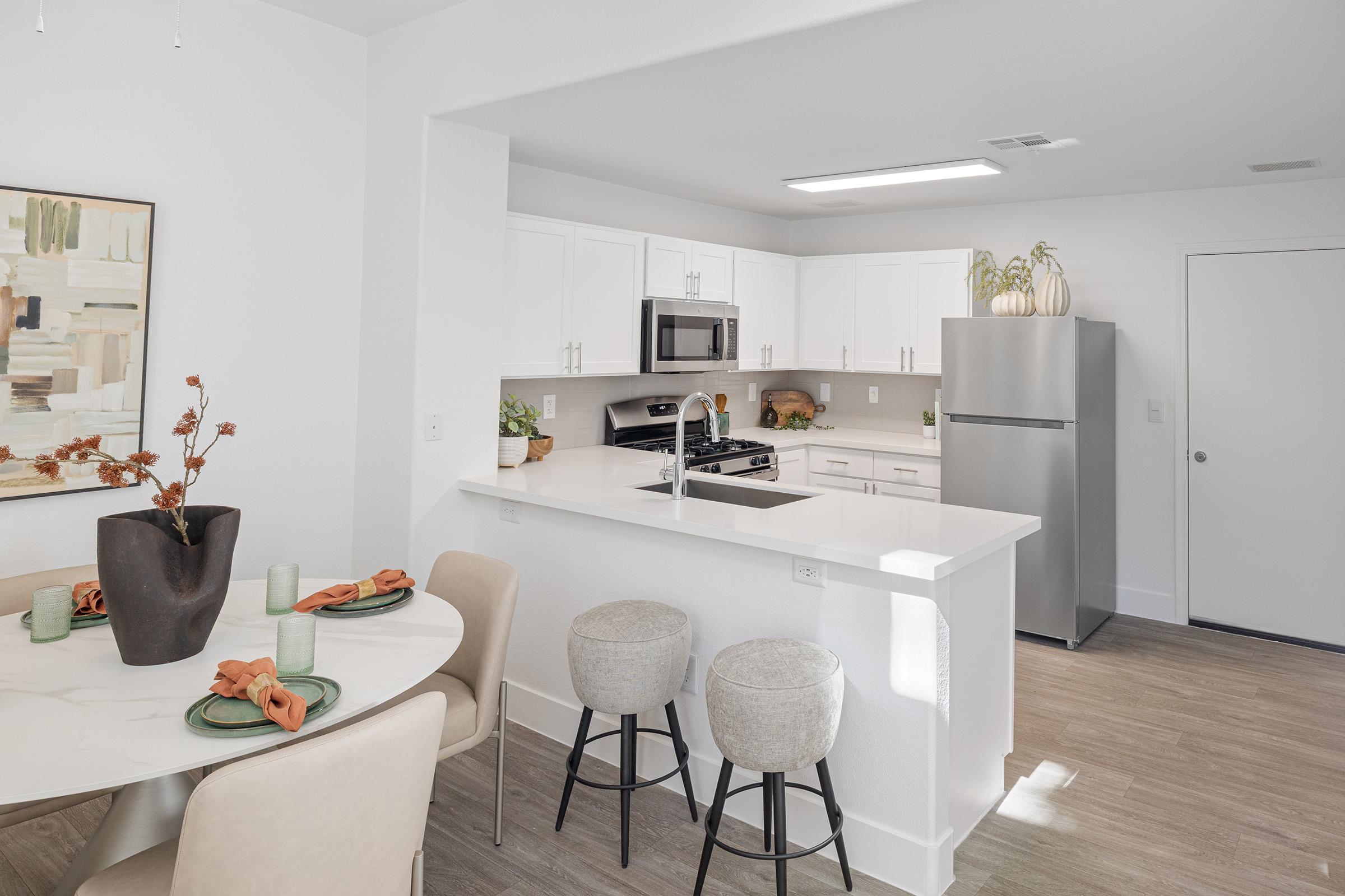Modern kitchen interior featuring a white color palette, stainless steel appliances, and a spacious countertop. A dining table with decorative elements is positioned nearby, along with comfortable bar stools. The space is well-lit and features plants for a touch of greenery.
