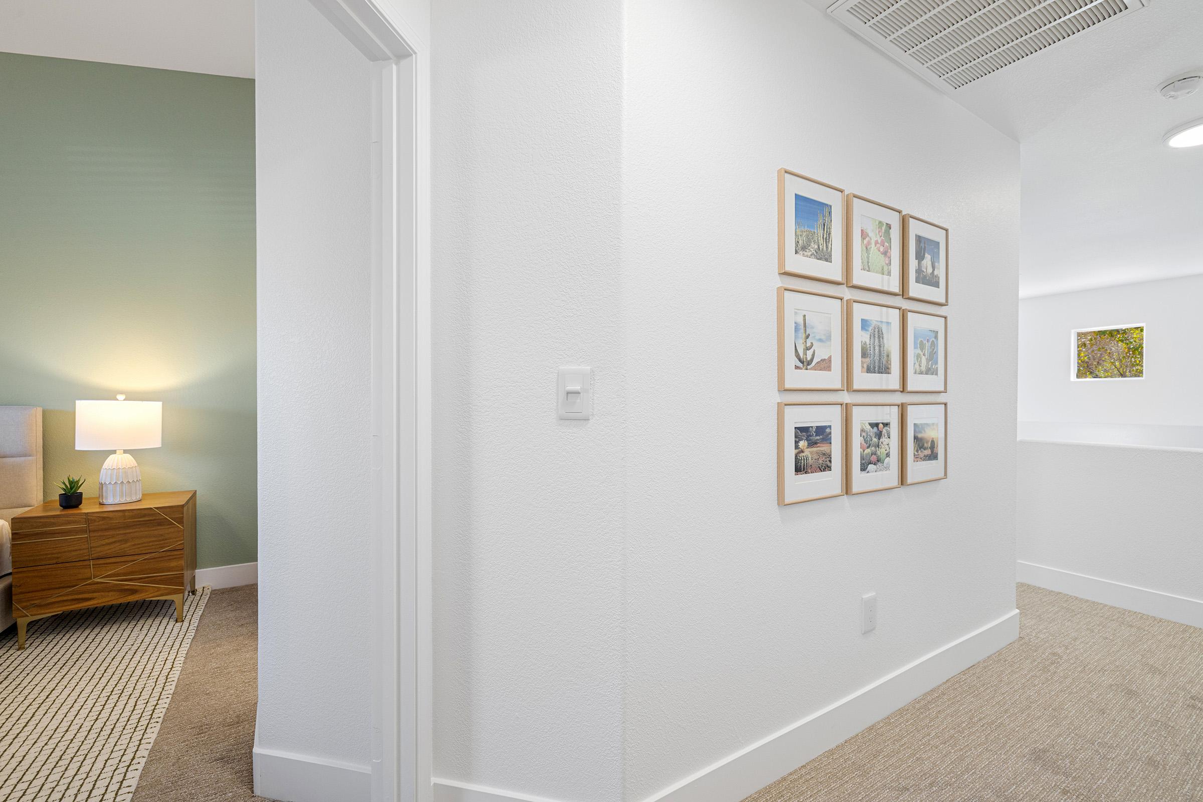A well-lit hallway with a light-colored wall, featuring a grid of framed photographs on the right. A cozy side table with a lamp is visible on the left, adjacent to a room with a green accent wall. The flooring is carpeted, and the overall decor is modern and minimalist.