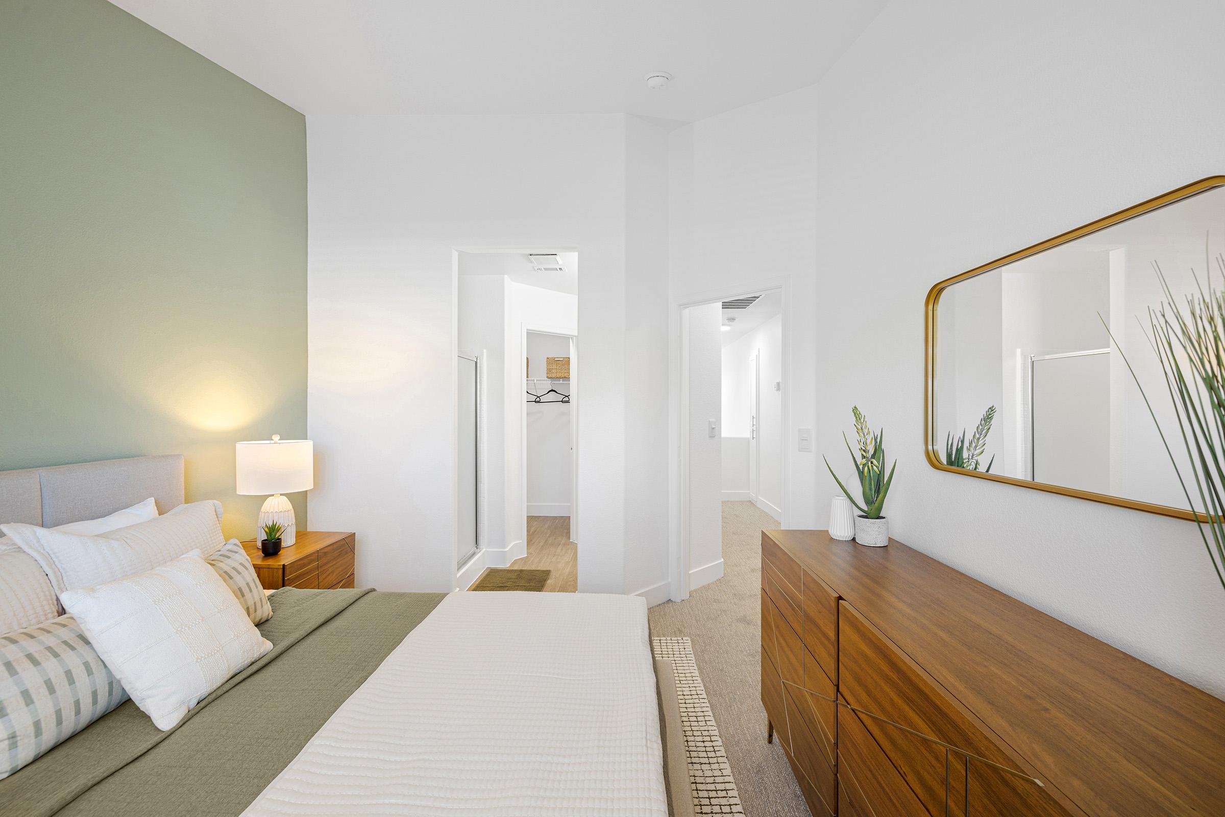 A modern bedroom featuring a light green accent wall, a queen-sized bed with white and green bedding, a bedside lamp on a wooden nightstand, and a stylish wooden dresser. Natural light fills the space, highlighting minimalistic decor and indoor plants for a fresh ambiance.