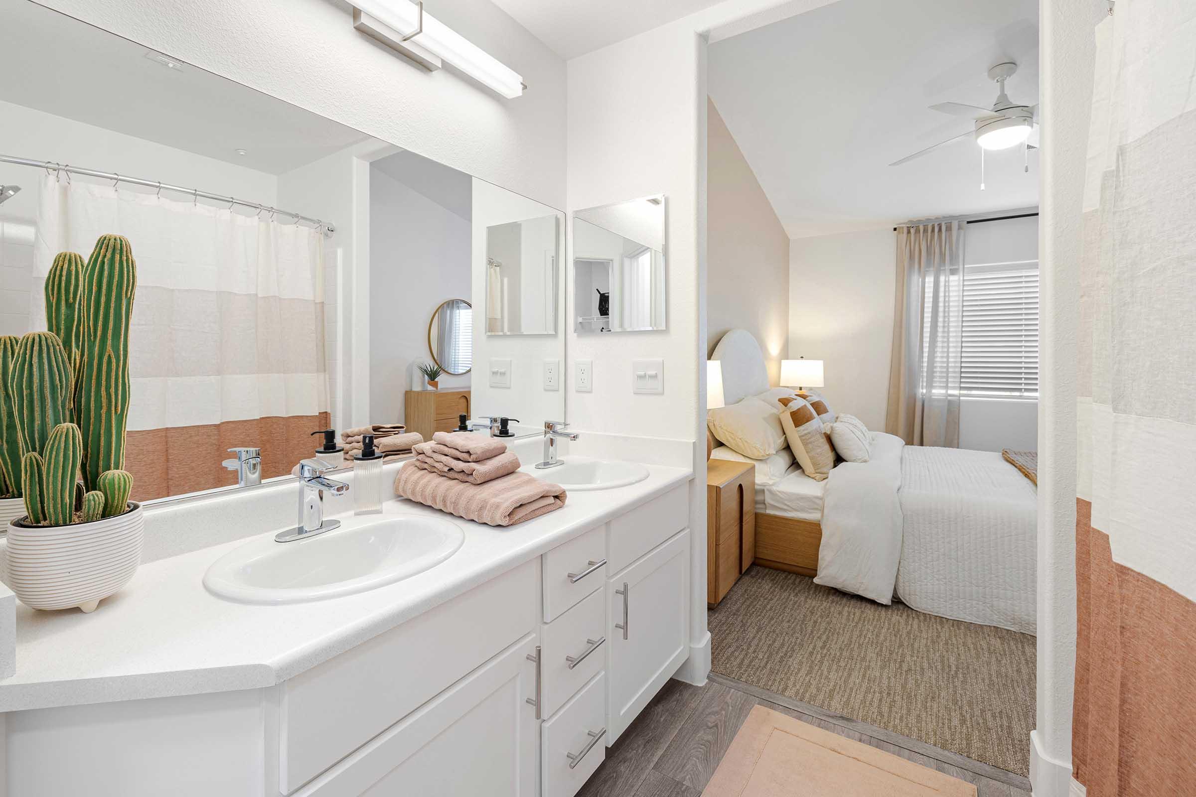 A modern bathroom featuring double sinks with a minimalist design. There are neatly stacked towels in earthy tones, a potted cactus on the counter, and a large mirror. Adjacent is a well-lit bedroom with a bed dressed in light-colored linens and a ceiling fan, creating a warm and inviting atmosphere.