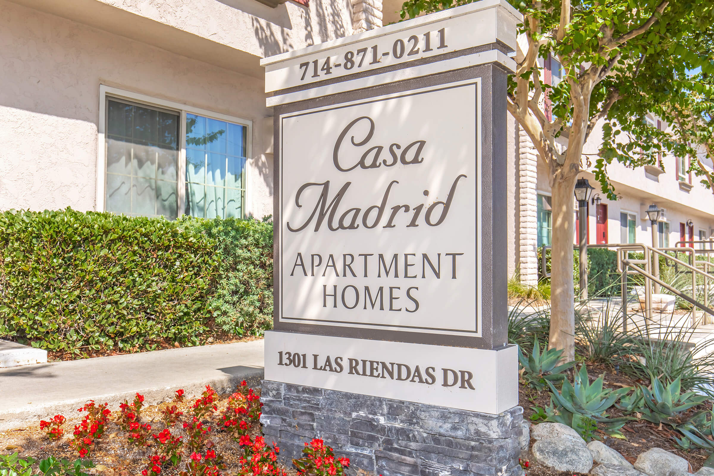 Sign for Casa Madrid Apartment Homes, located at 1301 Las Riendas Drive. The sign includes the phone number 714-871-0211, surrounded by greenery and flowers.