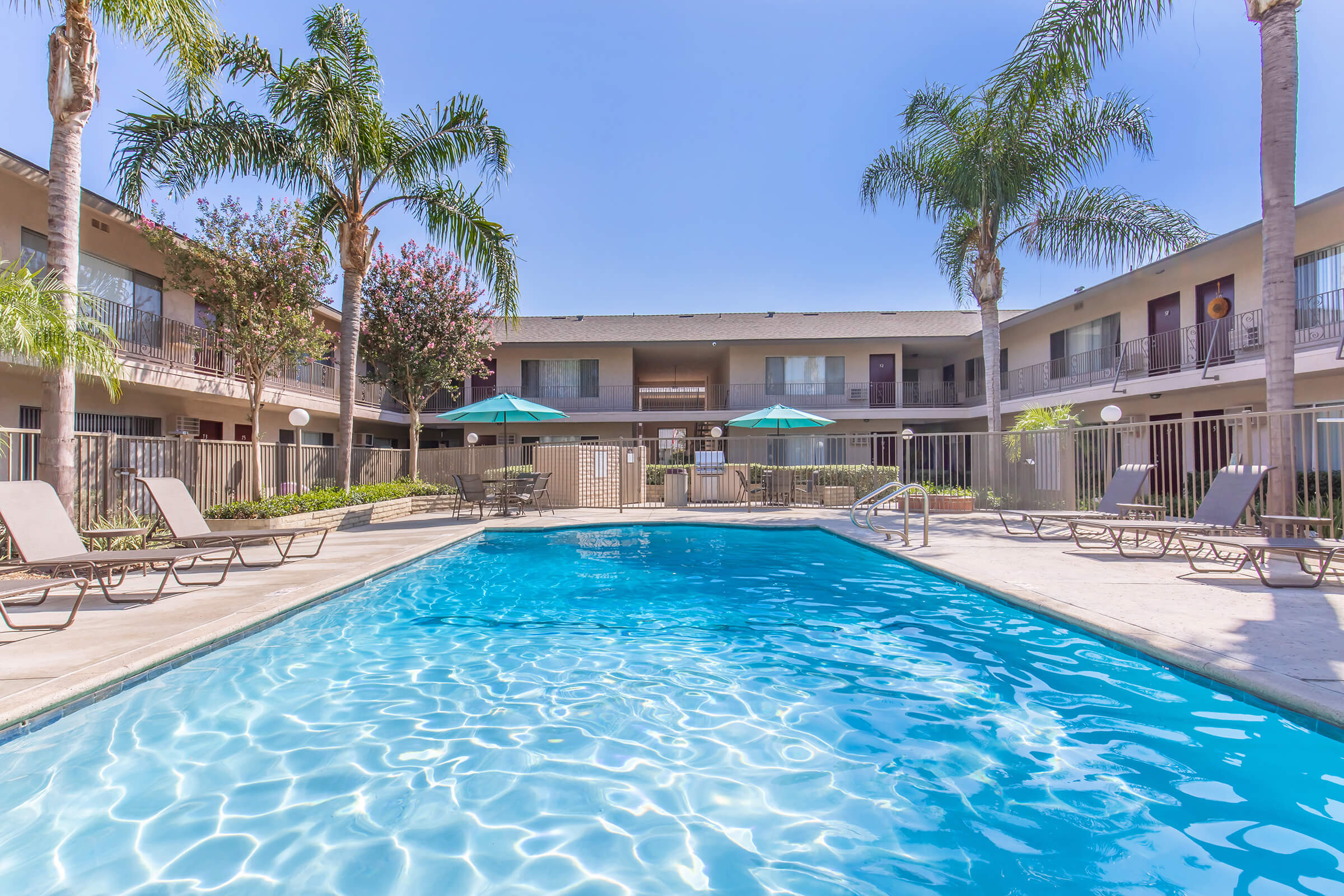 A clear blue swimming pool surrounded by lounge chairs, with green umbrellas providing shade. Palm trees and flowering plants are visible around the pool area, with two-story buildings in the background. The sky is bright and sunny, creating a relaxing atmosphere for guests.