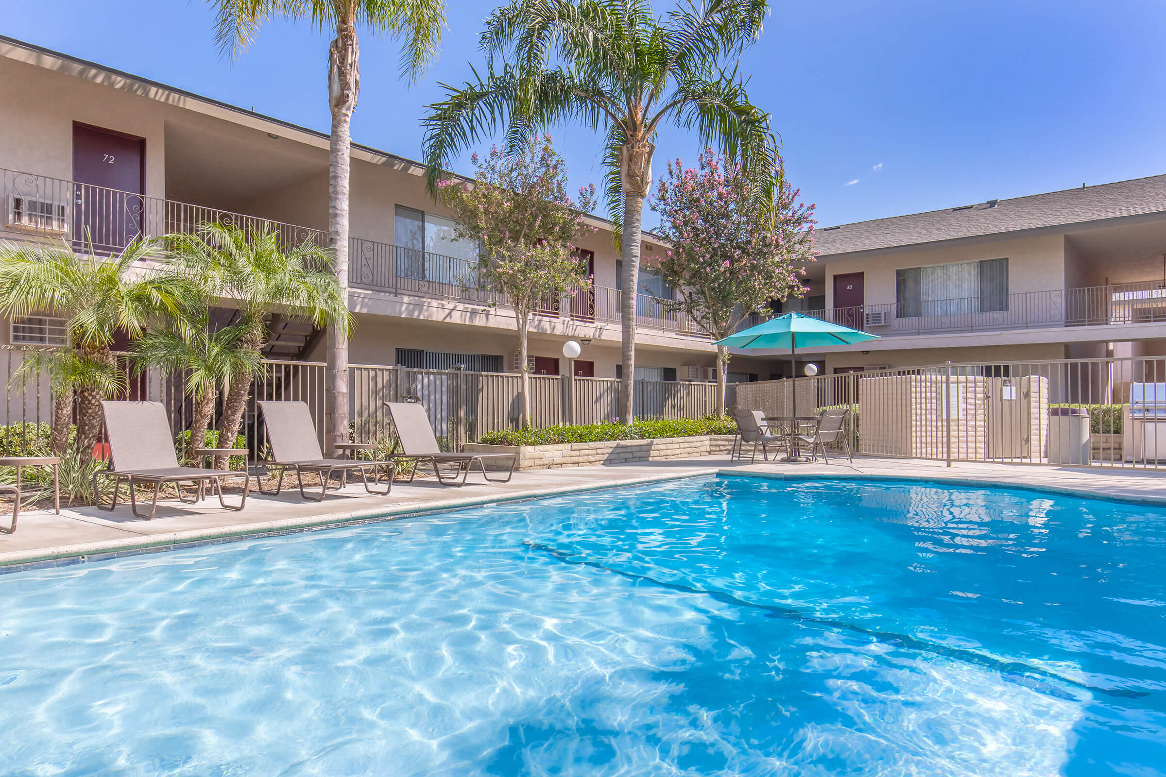 A sparkling blue swimming pool surrounded by lounge chairs, with palm trees and flowering plants nearby. In the background, there's a two-story building with balconies and doors. An umbrella-shaded table adds to the inviting atmosphere on a sunny day.