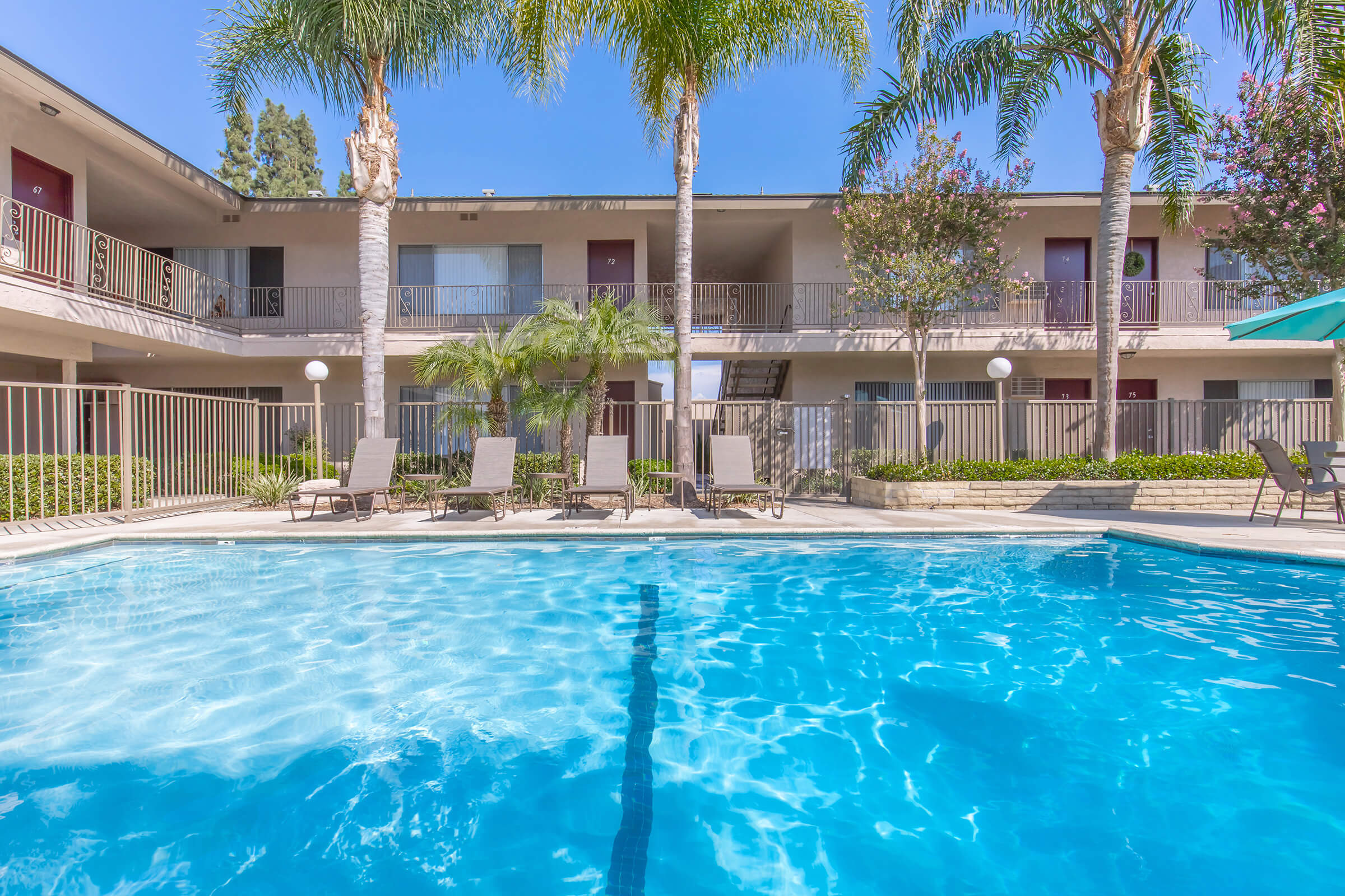 A clear swimming pool surrounded by lounge chairs and palm trees, with a multi-story building in the background featuring balconies and greenery. The scene is bright and inviting, perfect for relaxation.
