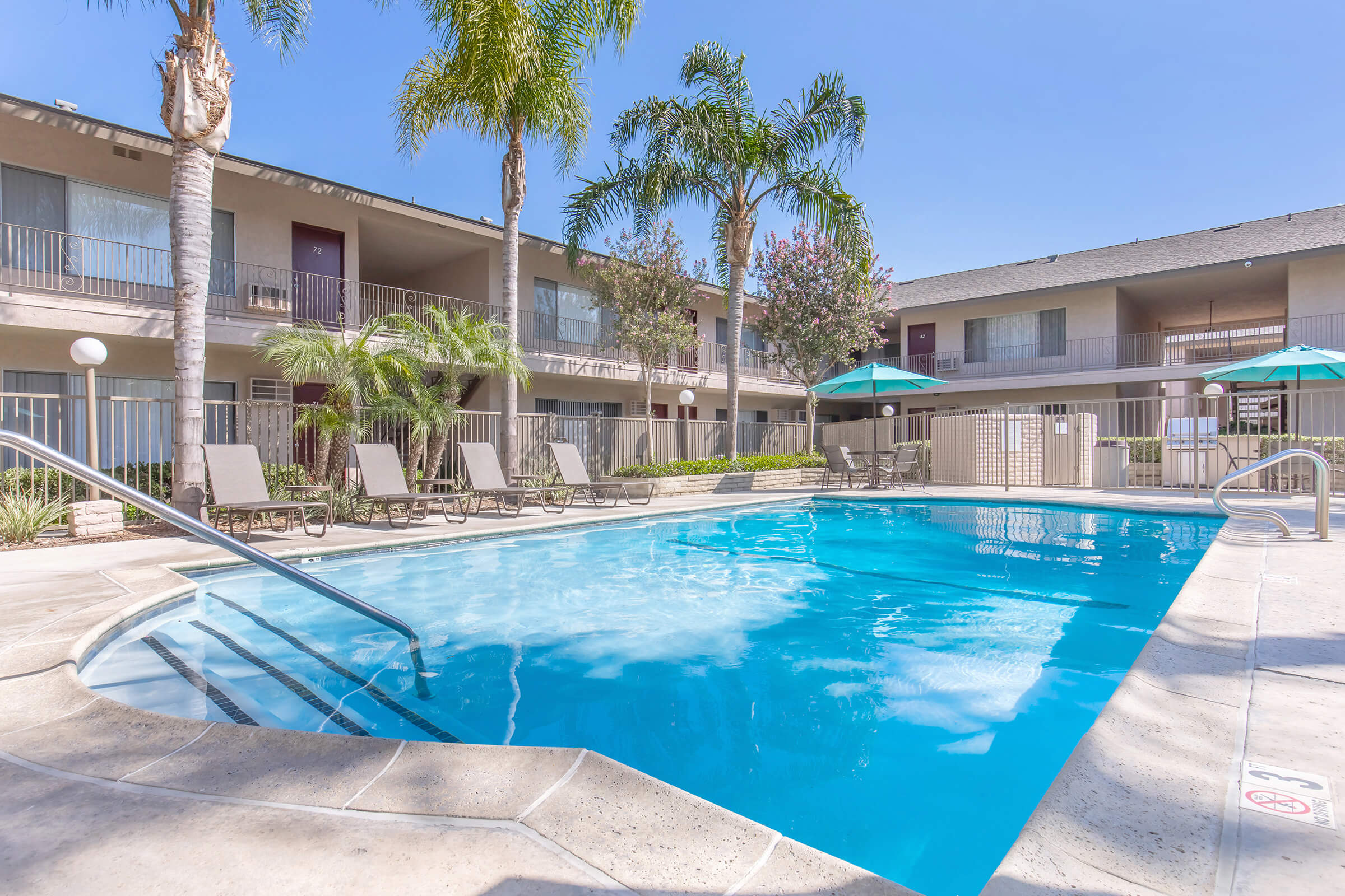 A clear blue swimming pool surrounded by lounge chairs, palm trees, and shaded umbrellas. Two-story buildings with balconies are visible in the background against a clear sky.