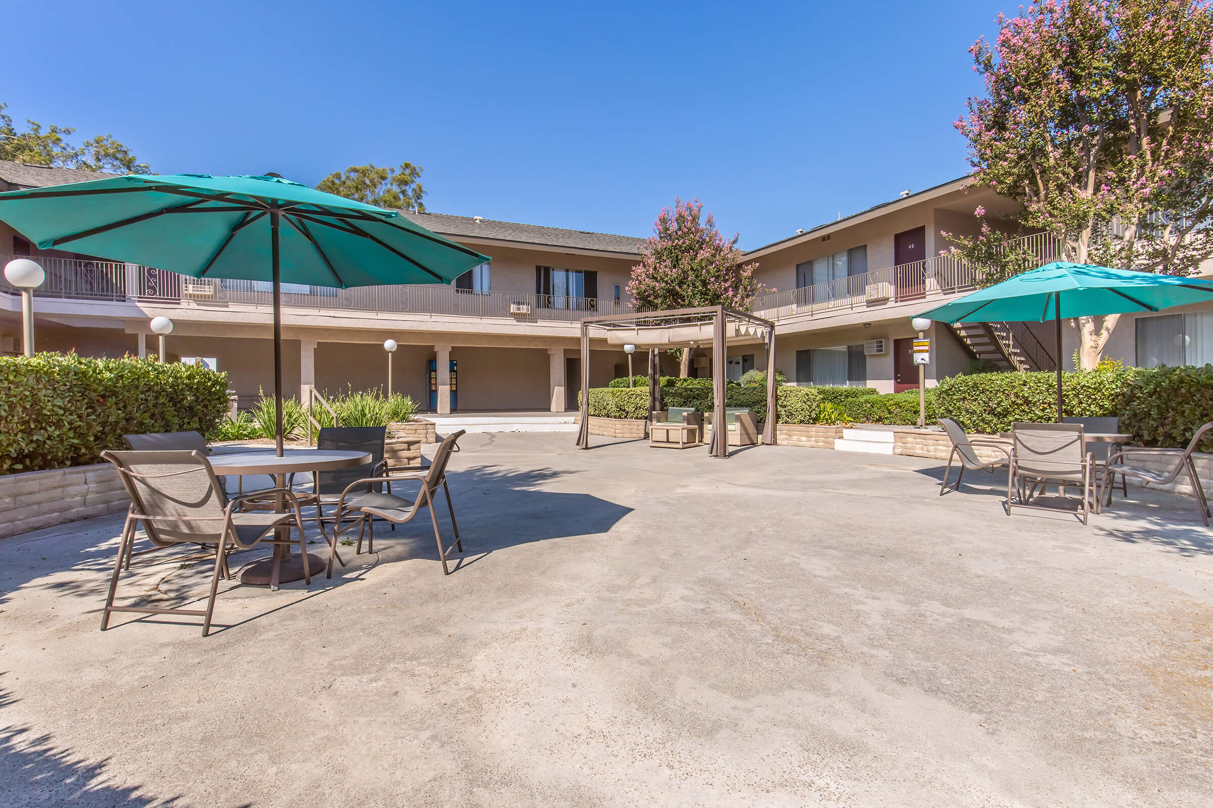 A sunny courtyard area featuring green umbrellas over tables and chairs, surrounded by lush greenery and flowering bushes. In the background, two-story buildings with balconies are visible, creating a welcoming environment for relaxation and socializing. Clear blue skies add to the cheerful atmosphere.