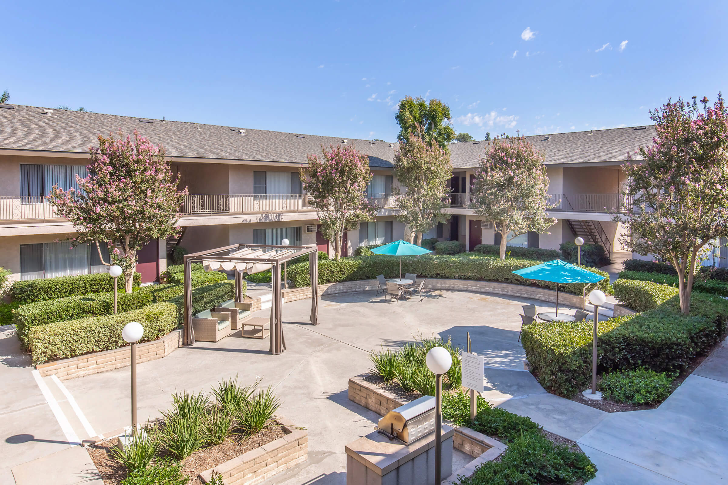 A view of a courtyard at a hotel featuring landscaped greenery, blooming trees, and seating areas. There are two umbrellas providing shade over tables and chairs. The architecture includes two-story buildings with balconies and open walkways, all set against a clear blue sky.