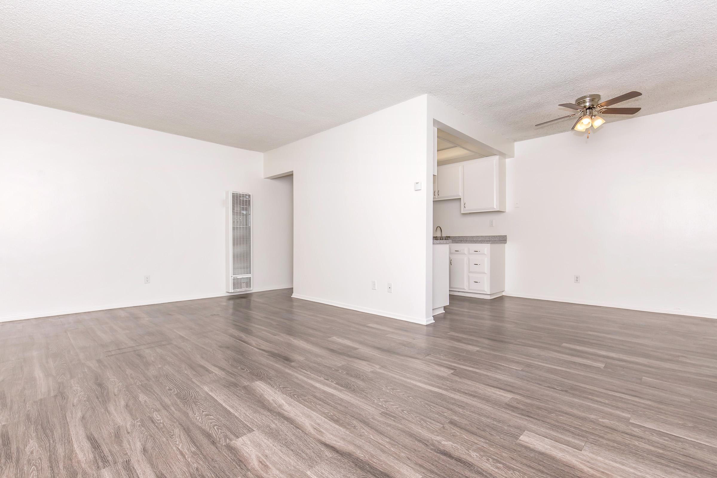 A spacious, empty living area featuring light-colored walls and modern laminate flooring. A ceiling fan is installed, and a doorway leads to a kitchen with white cabinetry visible. The room has a clean, minimalist design with ample natural light.