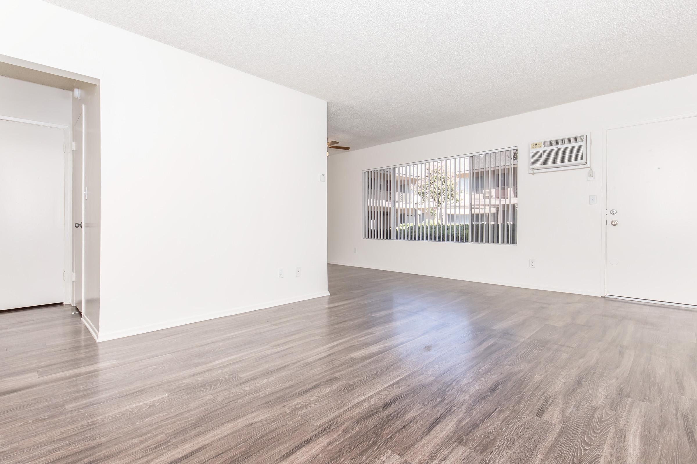A spacious, empty living room with light-colored walls and a textured ceiling. The floor features light wood-like laminate. A large window with security bars allows natural light in, and there is an air conditioning unit mounted on the wall. The entrance door is visible, along with an adjacent room entrance.