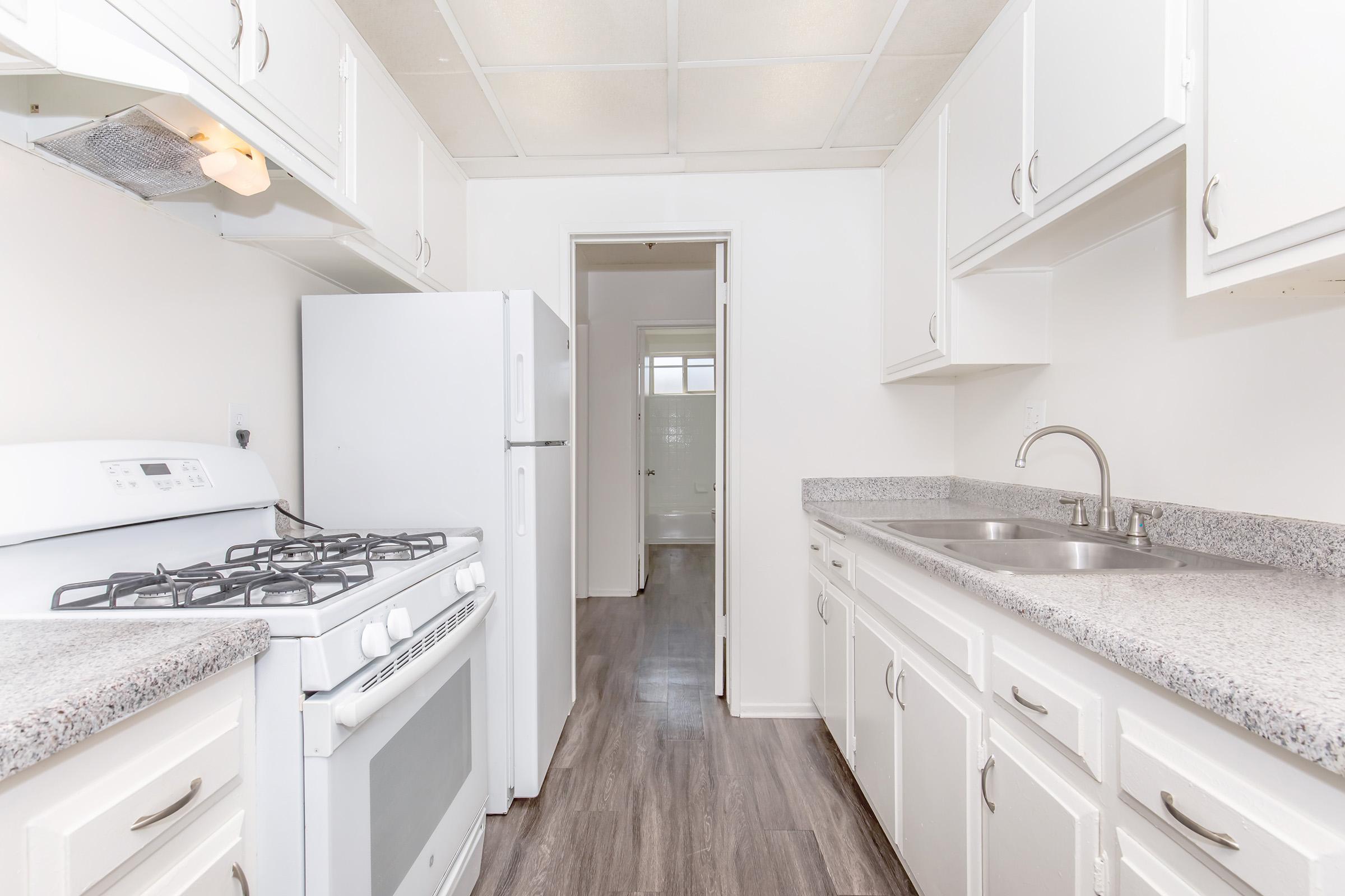 A clean, modern kitchen featuring white cabinets, a gas stove, and a double sink. The kitchen has a light color scheme with gray countertops. In the background, there is a hallway leading to another room, illuminated by natural light. The floor is made of light-colored wood or laminate.