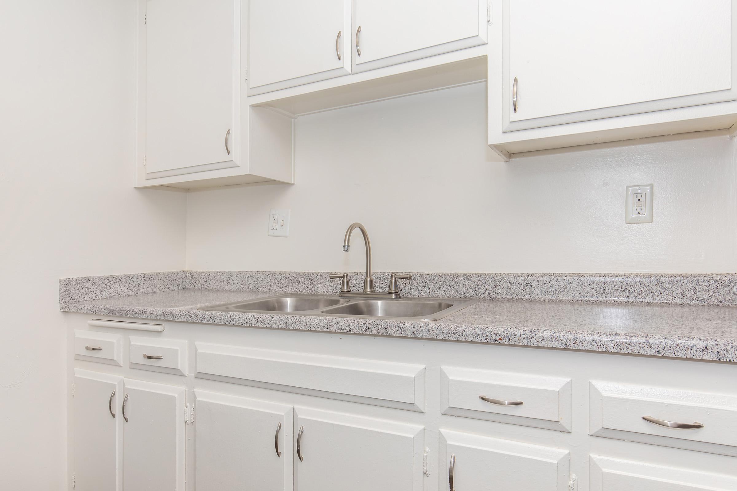 Modern kitchen featuring white cabinets, a grey speckled countertop, and a double sink with a sleek faucet. The walls are painted white, and the overall aesthetic is clean and minimalist.