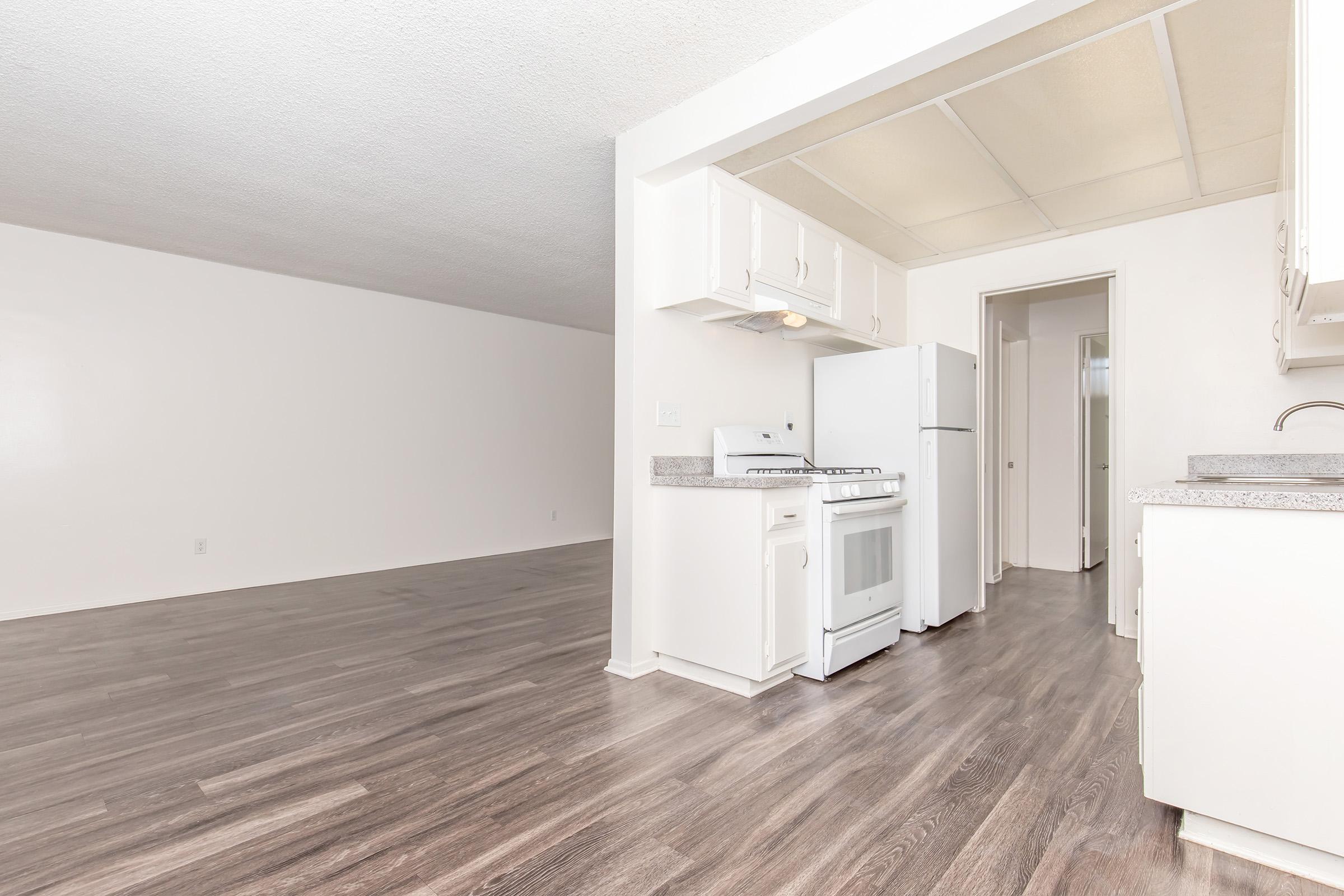 A spacious open-concept area featuring a kitchen with white cabinetry, a refrigerator, and a stove. The hardwood-like flooring extends into a large, empty living space with neutral-colored walls, creating a bright and airy atmosphere.