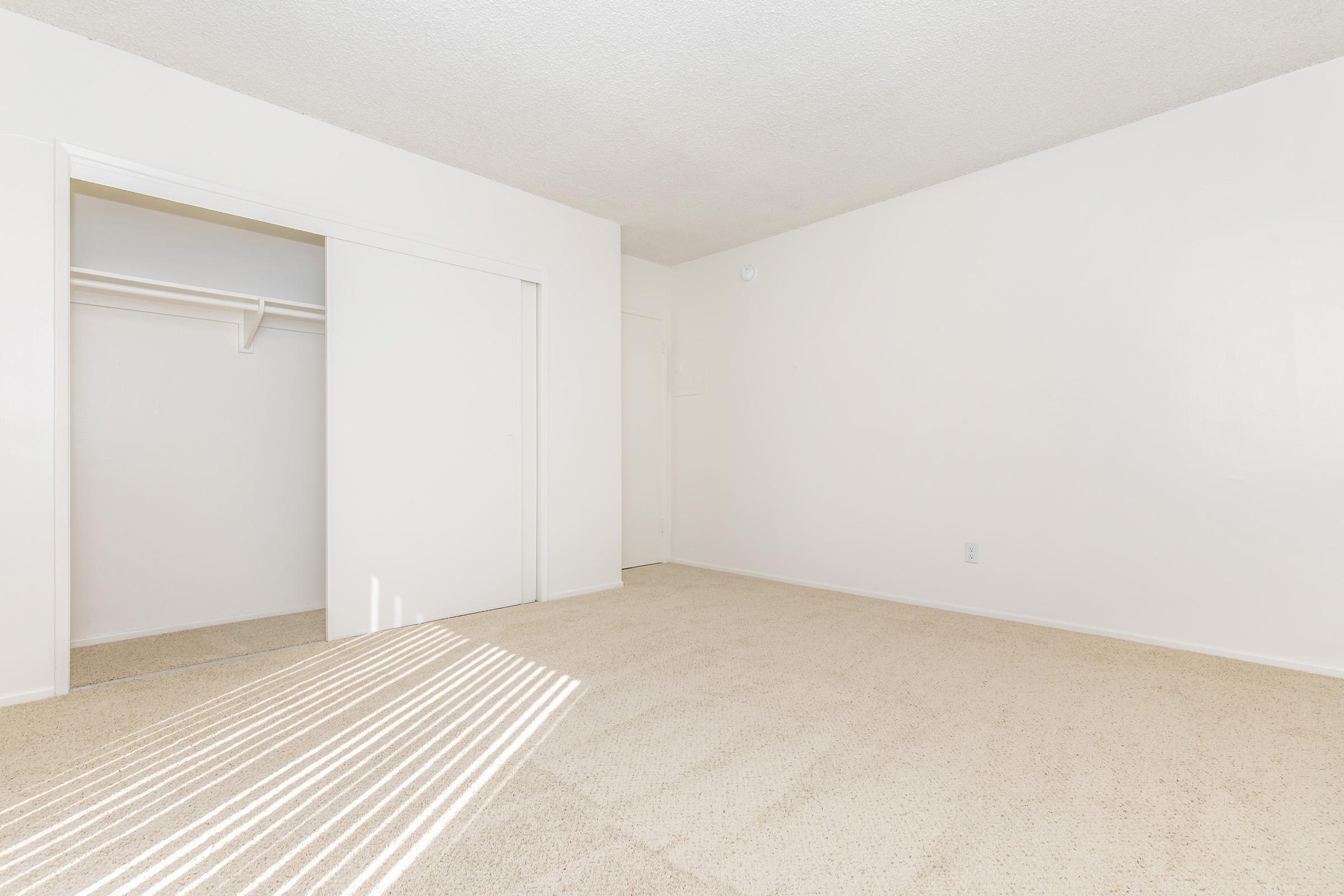 A spacious, empty room with beige carpet and white walls. There is a closet with sliding doors on the left side and a doorway leading to another room. Sunlight casts shadows on the floor from a window, suggesting a bright atmosphere. The overall feel is clean and uncluttered, ideal for personalization.