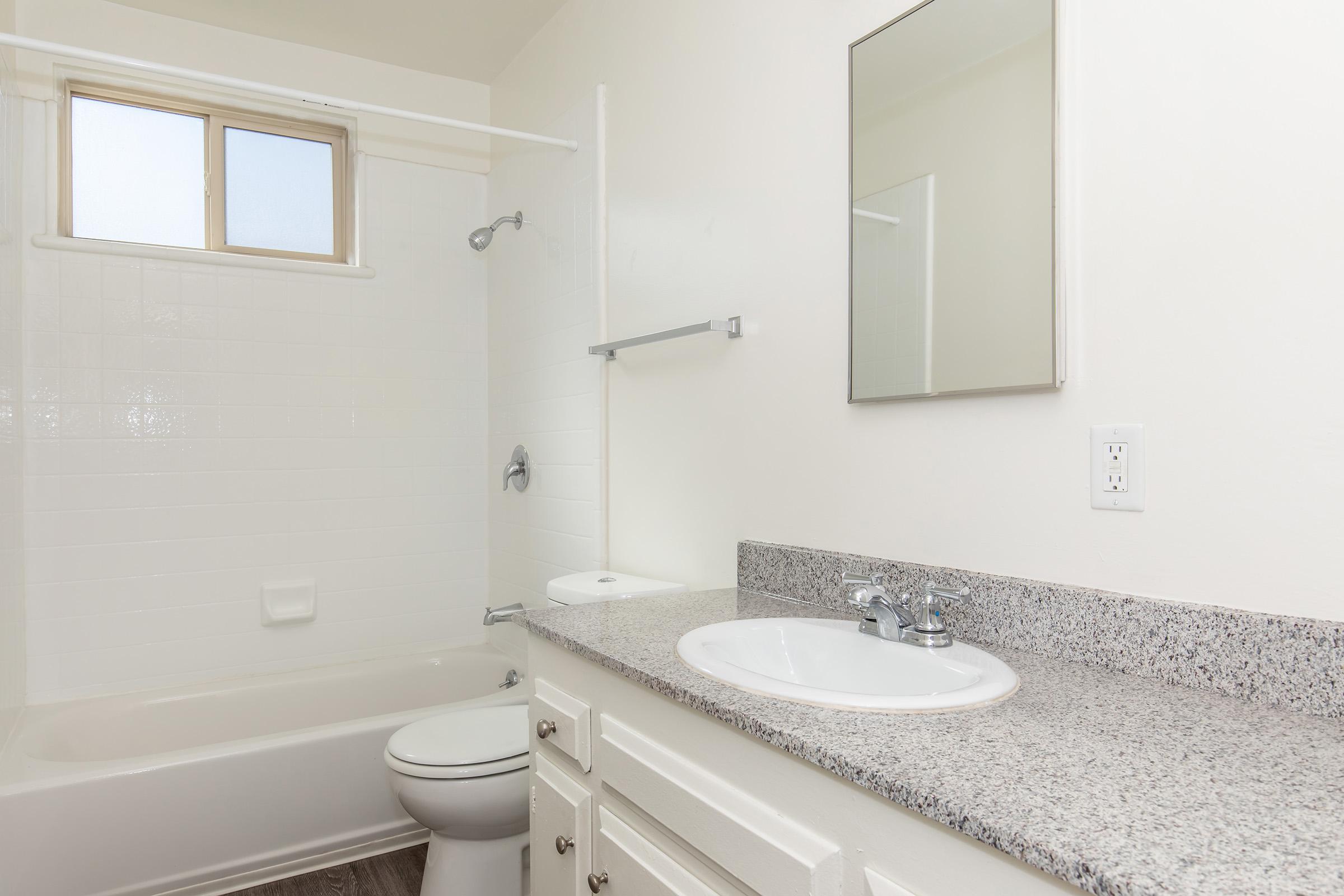 A clean and simple bathroom featuring a bathtub with a shower, a mirror above a white sink with a granite countertop, and light-colored walls. A small window provides natural light, and there is a toilet next to the sink. The overall design is minimalistic and modern.