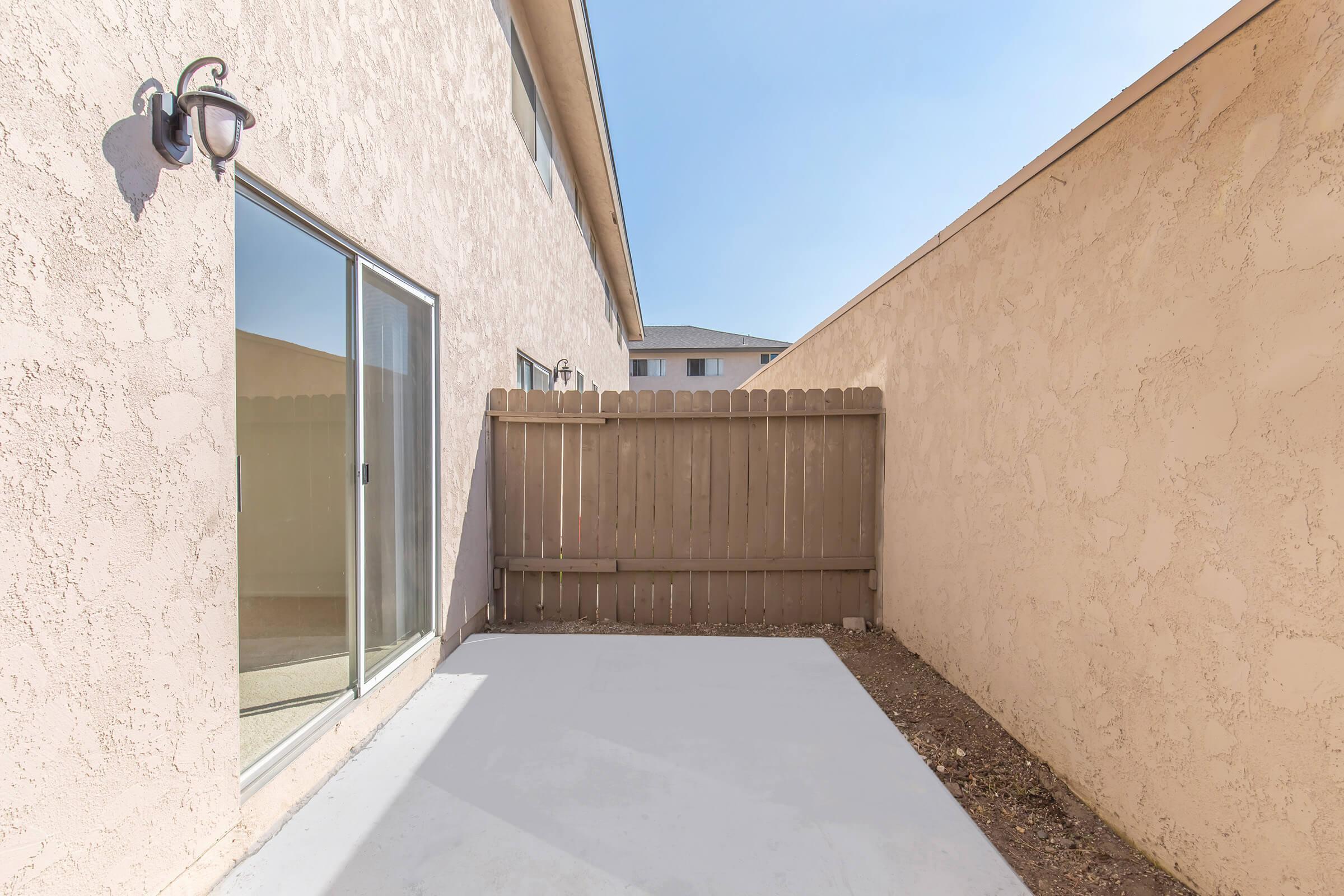 A narrow outdoor space with a concrete surface, featuring a wooden fence on one side and a wall on the other. The area is surrounded by beige-colored walls and includes a large glass door leading to an indoor area, with clear blue skies visible above.