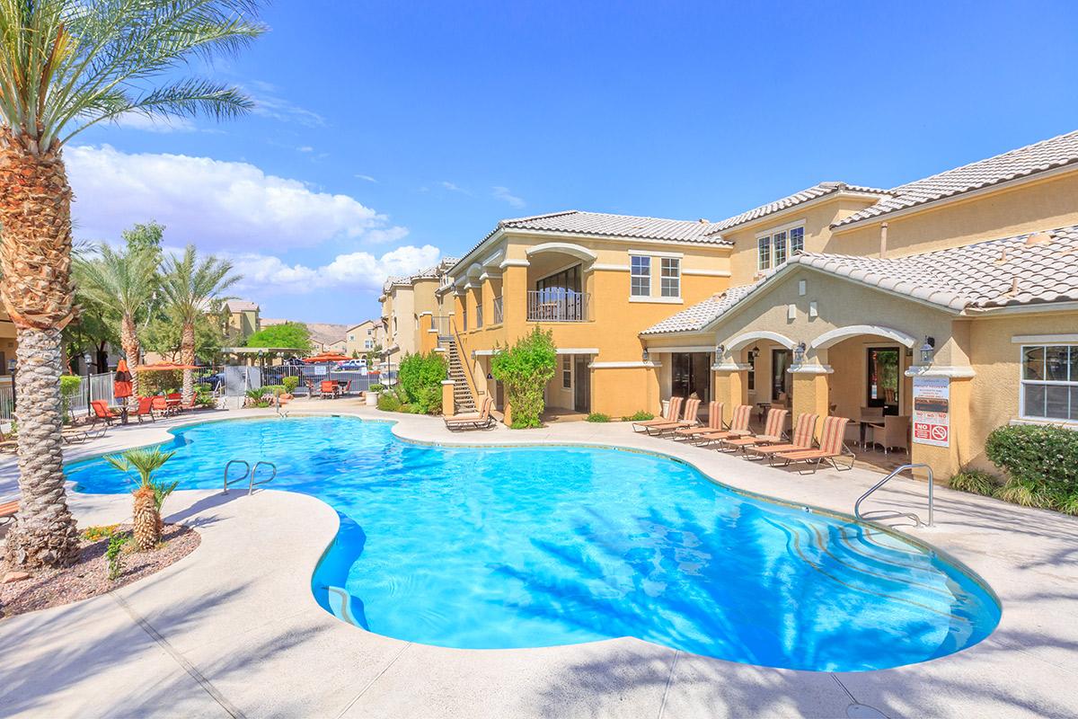 A resort-style swimming pool surrounded by palm trees and lounge chairs. The pool features a curved shape and is located next to a modern yellow building with balconies. Bright blue skies and fluffy white clouds complete the sunny ambiance, creating a relaxing outdoor setting.