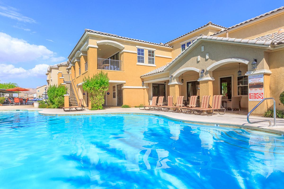 A clear blue swimming pool surrounded by lounge chairs, with a tan-colored building in the background. The sky is bright with a few fluffy clouds. The setting appears inviting and sunny, suggesting a relaxing atmosphere by the poolside.