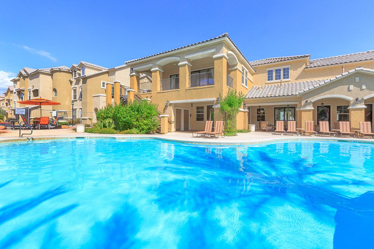 A sunny outdoor scene featuring a swimming pool surrounded by lounge chairs and lush greenery. In the background, a multi-story building with a tan exterior and balconies overlooks the pool area, set against a clear blue sky. A few umbrellas and patio furniture are visible near the water.
