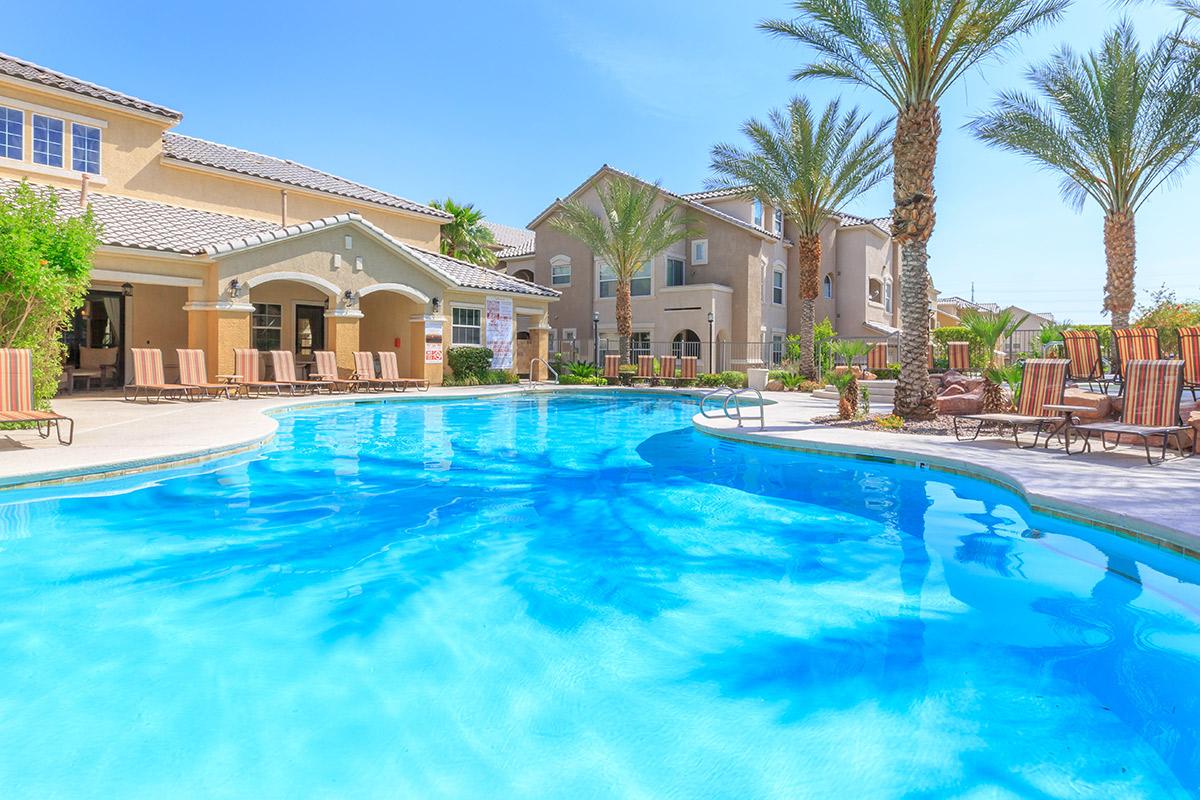 A bright, sunny pool area surrounded by palm trees and lounge chairs, with residential buildings in the background. The water is clear and inviting, reflecting the blue sky overhead. The scene conveys a relaxing, vacation-like atmosphere.