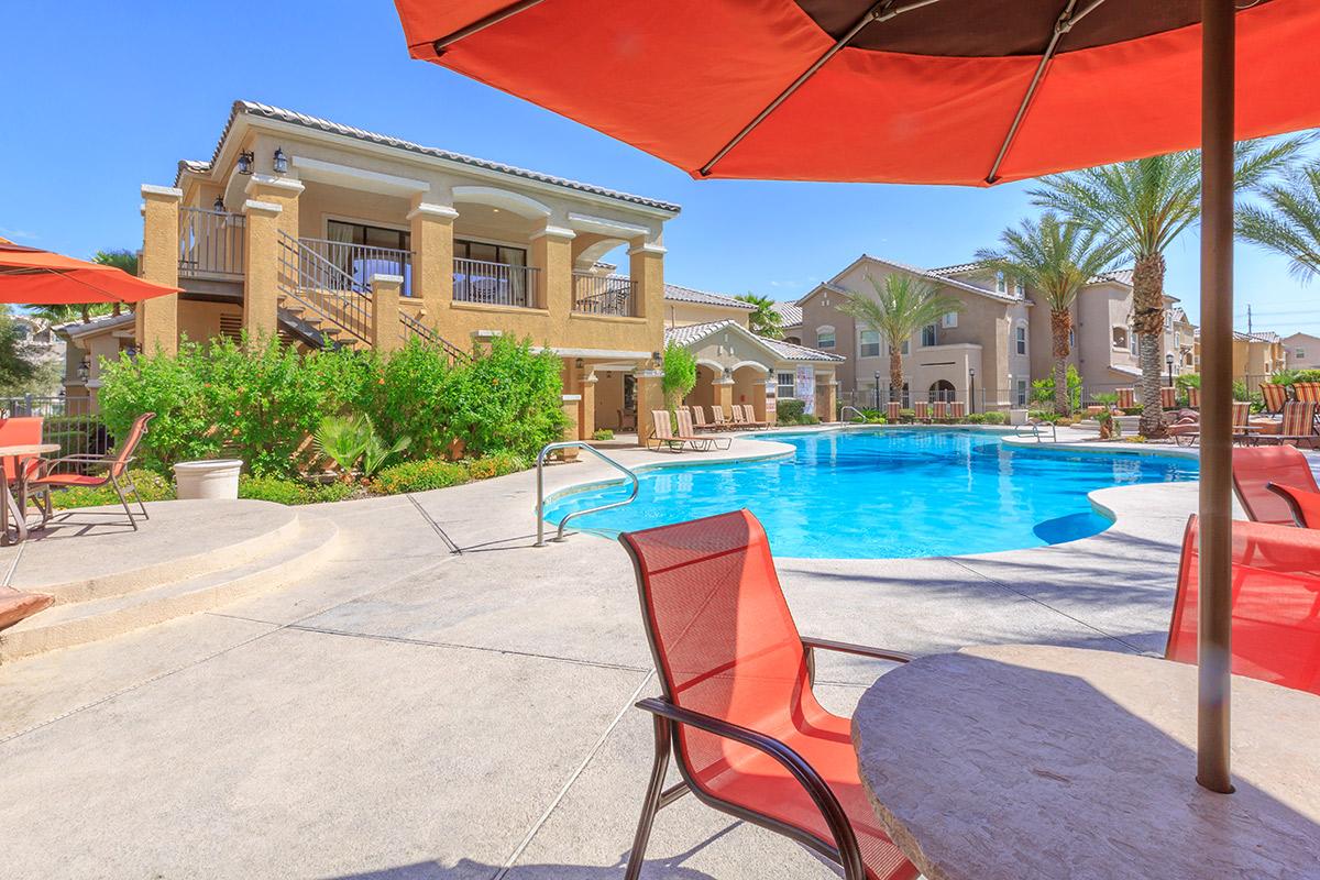 A sunny pool area featuring a refreshing blue swimming pool surrounded by lounge chairs, vibrant orange umbrellas, and green landscaping. In the background, several elegant multi-story buildings are visible, creating a relaxing outdoor environment perfect for leisure and relaxation.
