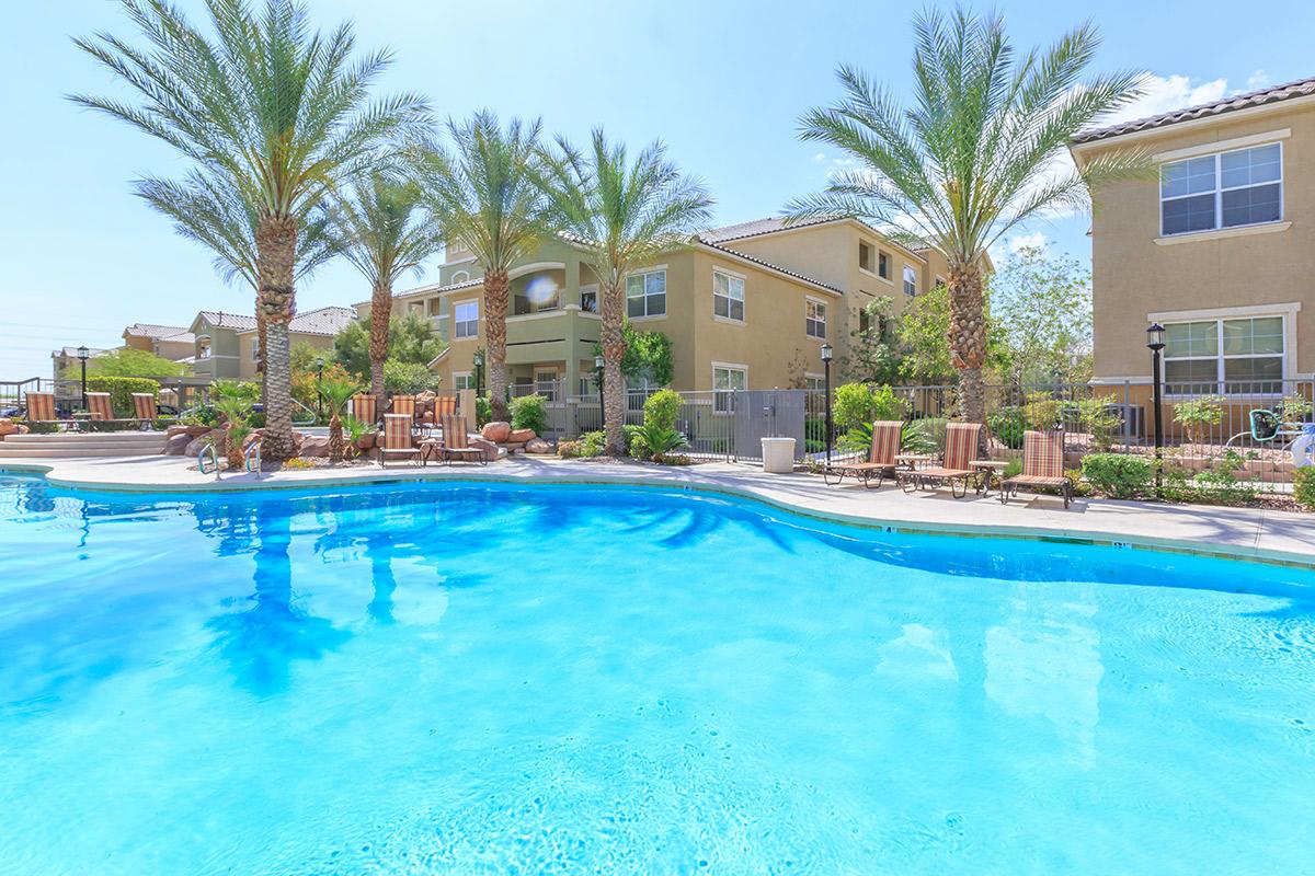 A clear blue swimming pool surrounded by palm trees and lounge chairs, with residential buildings in the background. Bright sunlight enhances the inviting atmosphere, creating a relaxing outdoor space ideal for leisure and recreation.