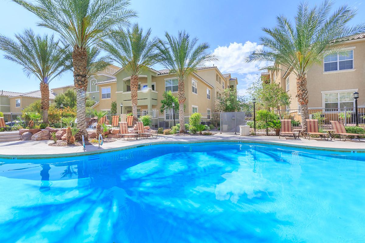A bright outdoor swimming pool surrounded by palm trees and lounge chairs, with beige apartment buildings in the background. The sky is clear and blue, creating a serene and inviting atmosphere.
